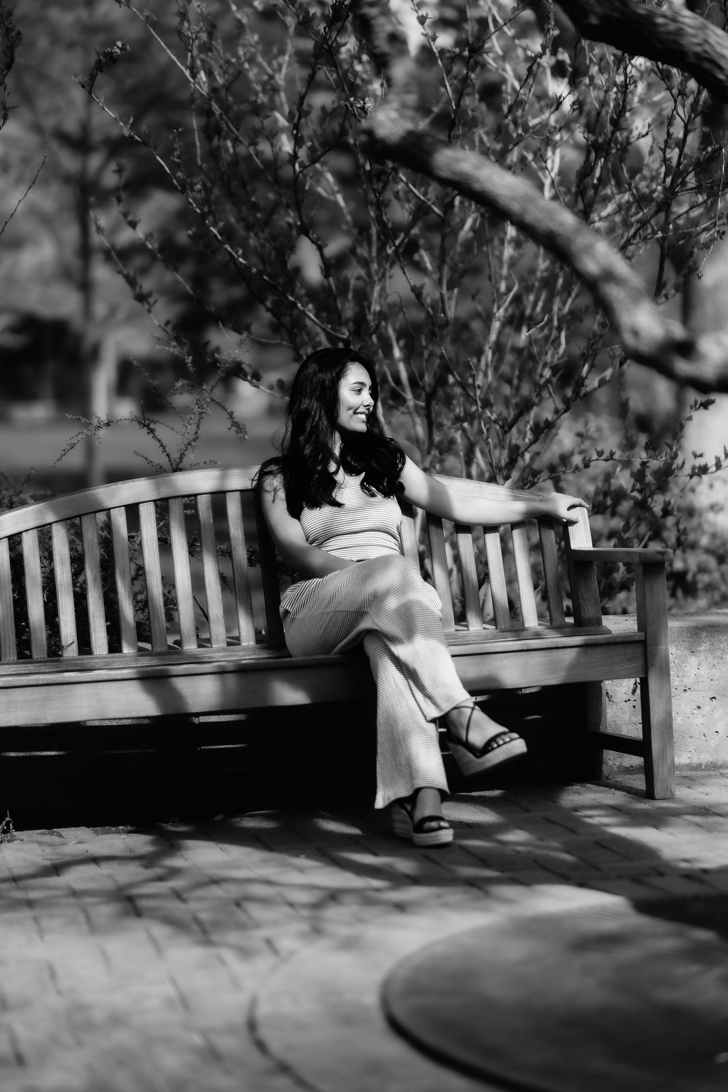 A monochrome candid shot of someone sitting casually on a wooden park bench surrounded by foliage.