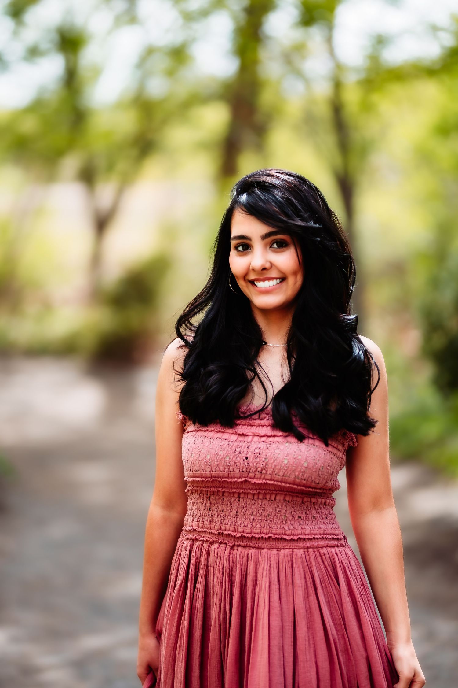 Portrait of someone in a dusty rose smocked dress posing outdoors with lush greenery in the background.