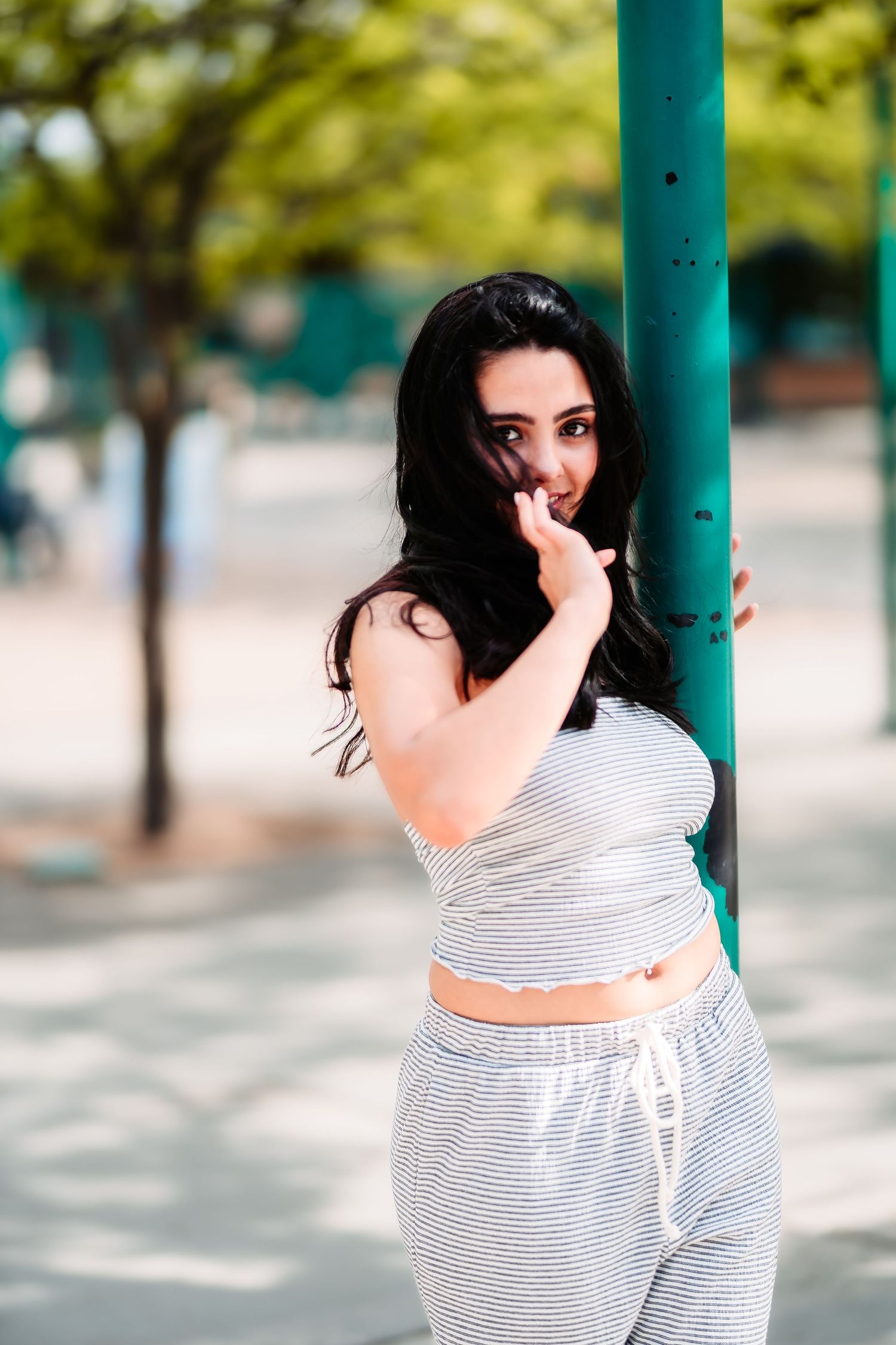A person in a striped crop top and matching pants playfully poses by a teal pole in an outdoor setting.