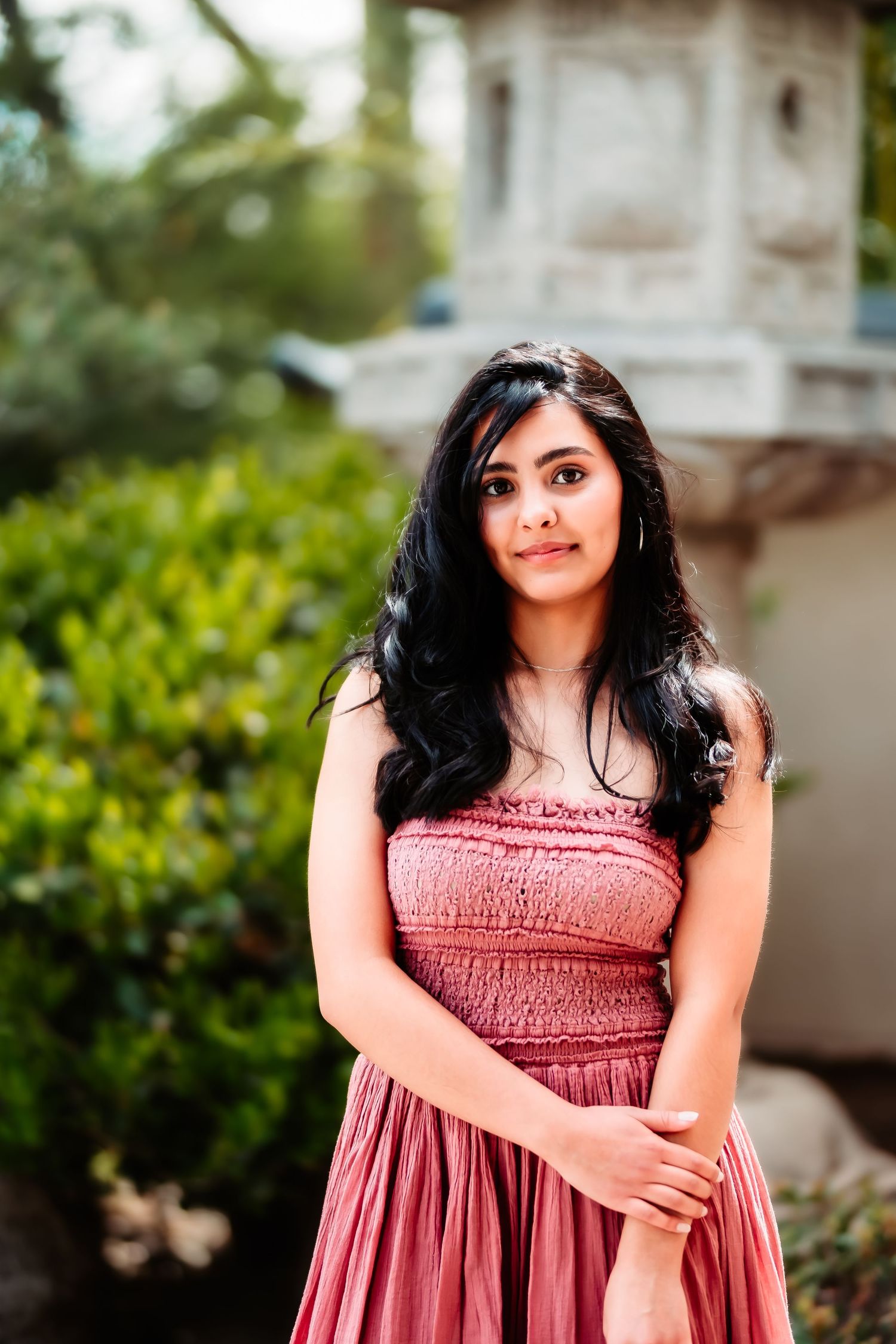 A woman in a pink smocked dress poses in a garden setting with greenery and architectural details in the background.
