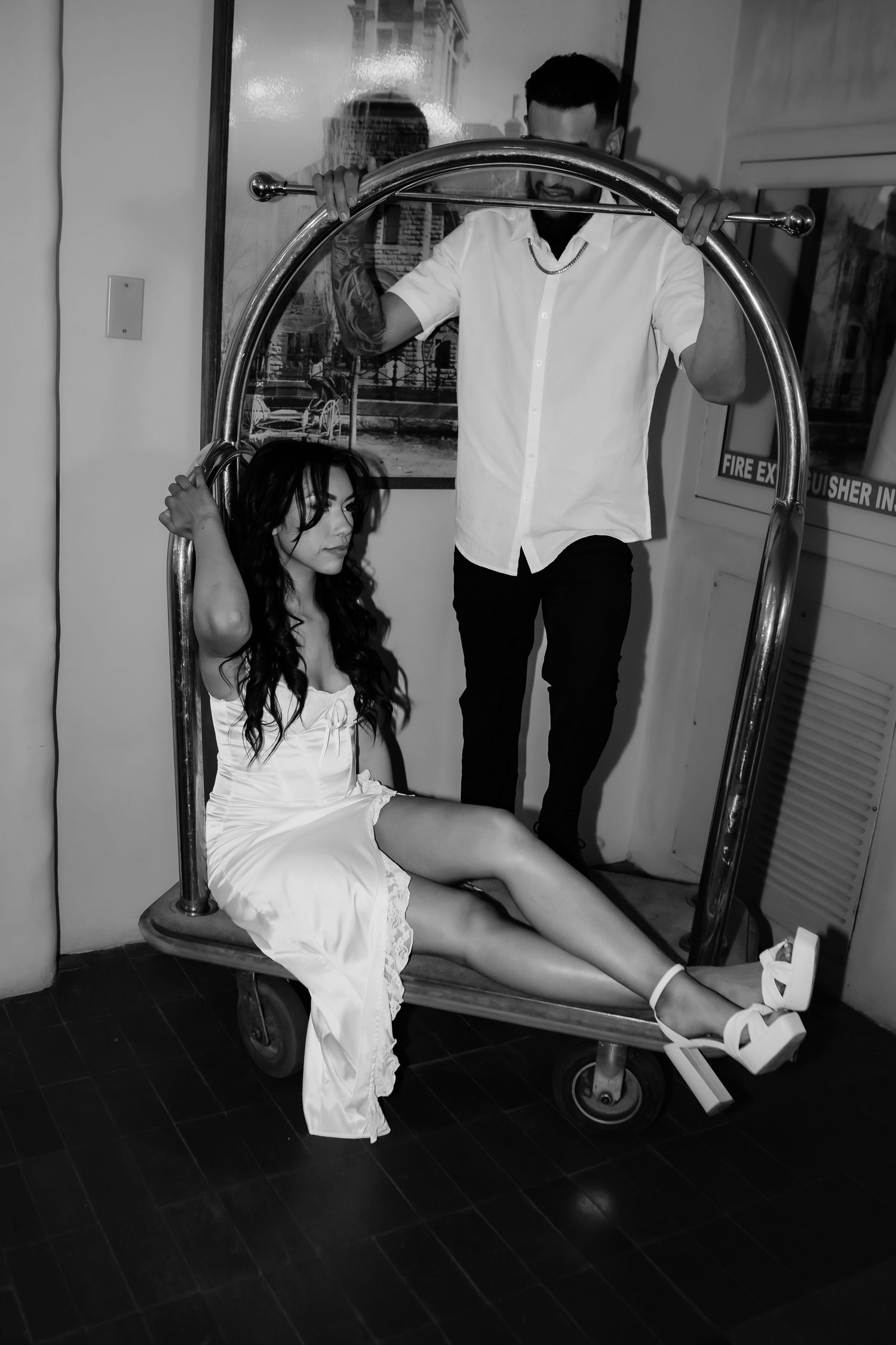 A couple poses dramatically near a vintage luggage cart in black and white photography.
