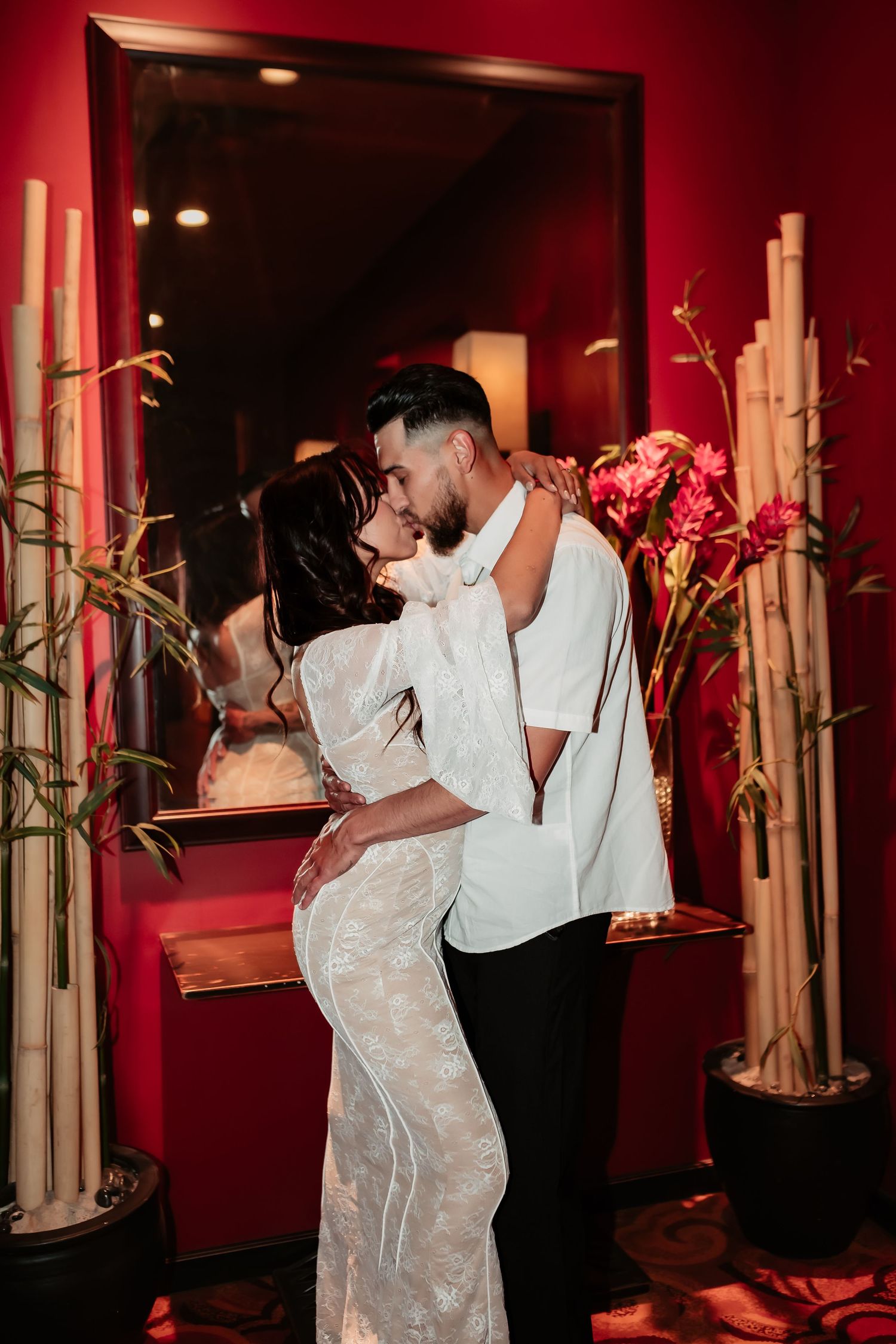 Romantic couple embrace in front of red wall with bamboo decor and mirror in an intimate setting.