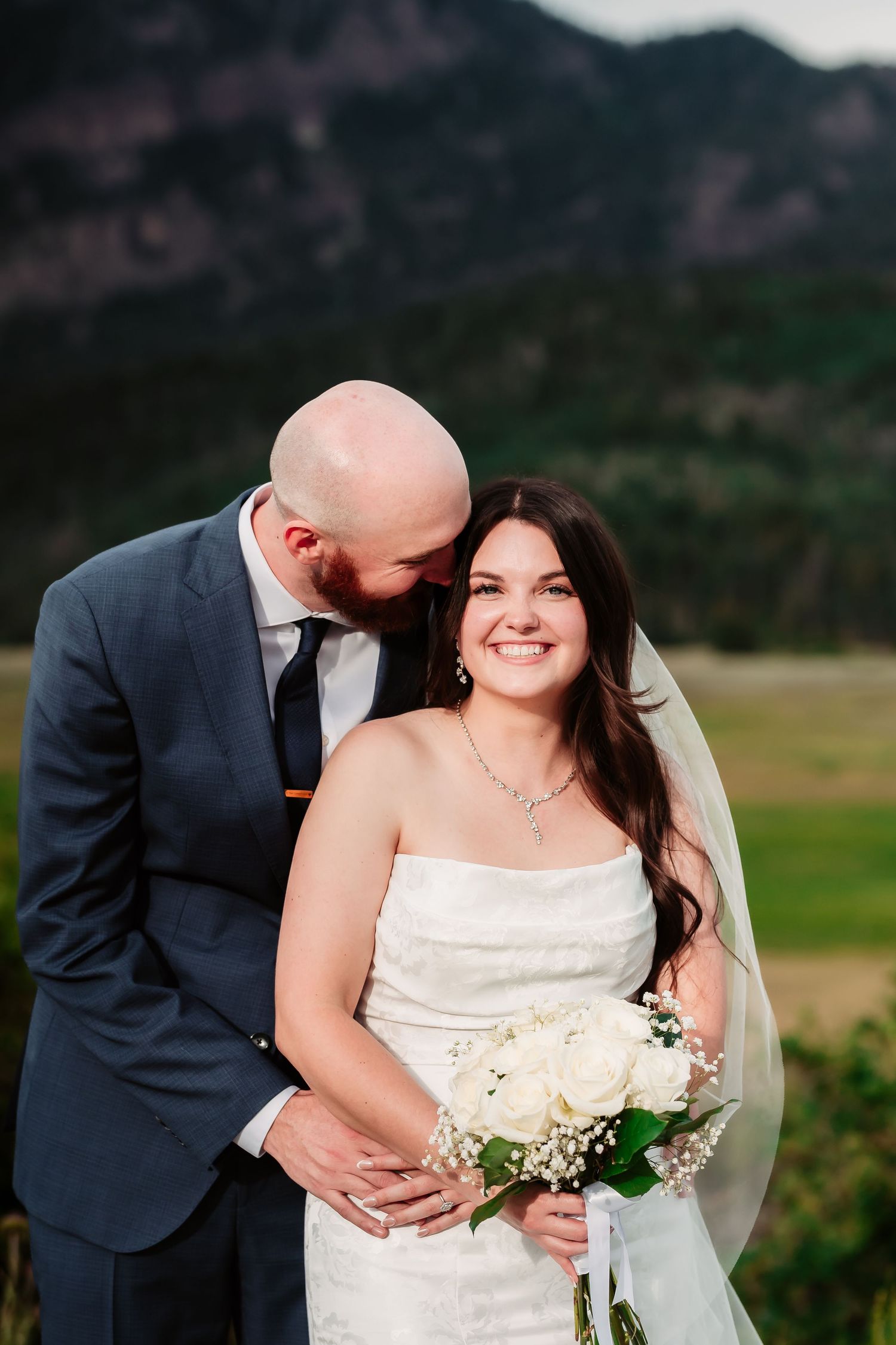 Newlyweds share an intimate moment against a mountain backdrop with the bride holding a delicate bouquet.