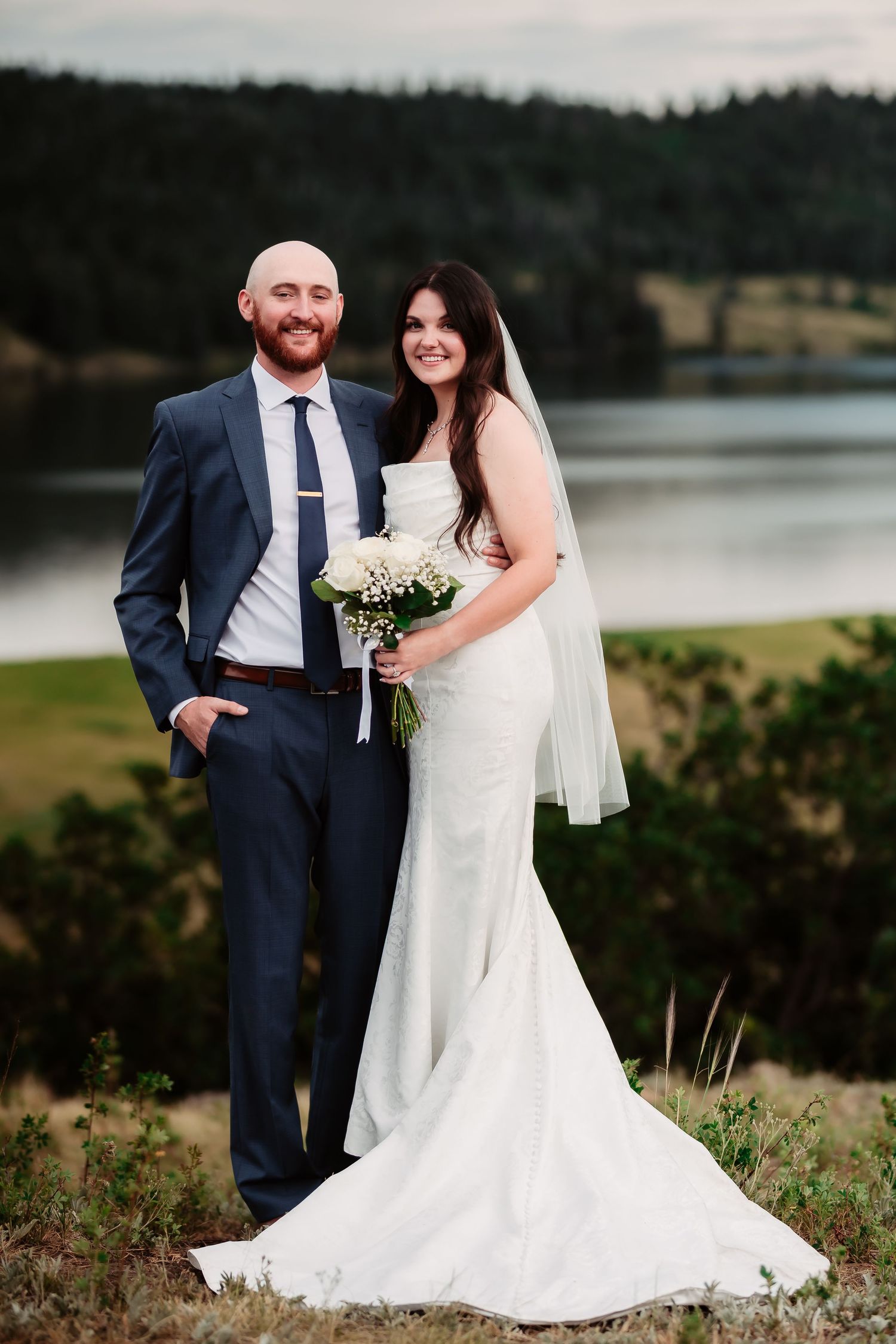 A outdoor wedding portrait with a beautiful lake and mountain backdrop on an overcast day.