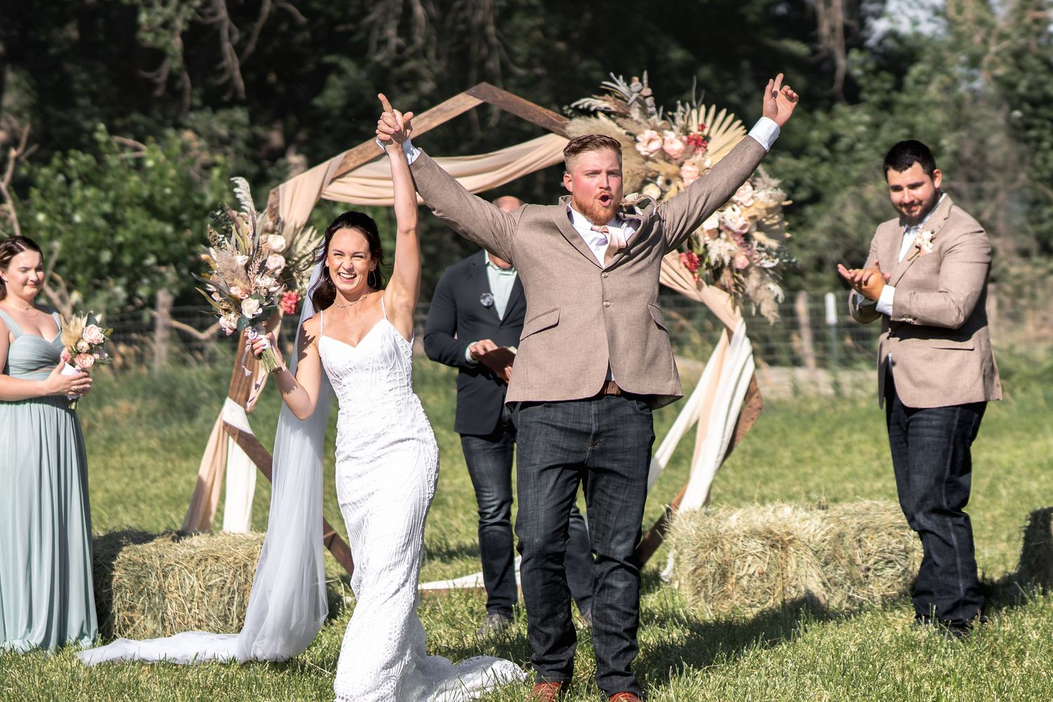 Newlyweds celebrate with raised arms under floral arch as guests applaud.
