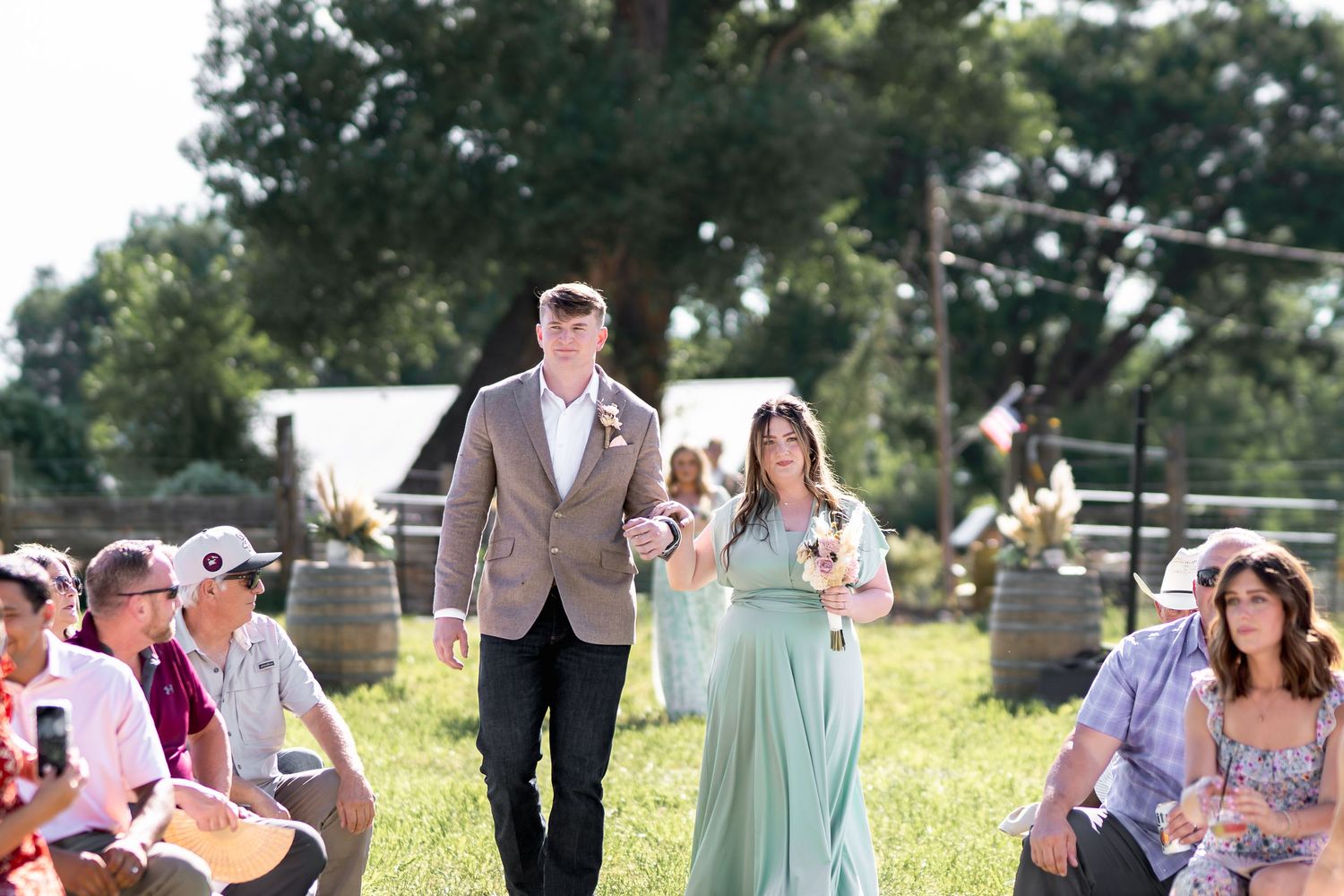 Wedding party members walk down a grassy aisle during an outdoor ceremony with guests seated on both sides.