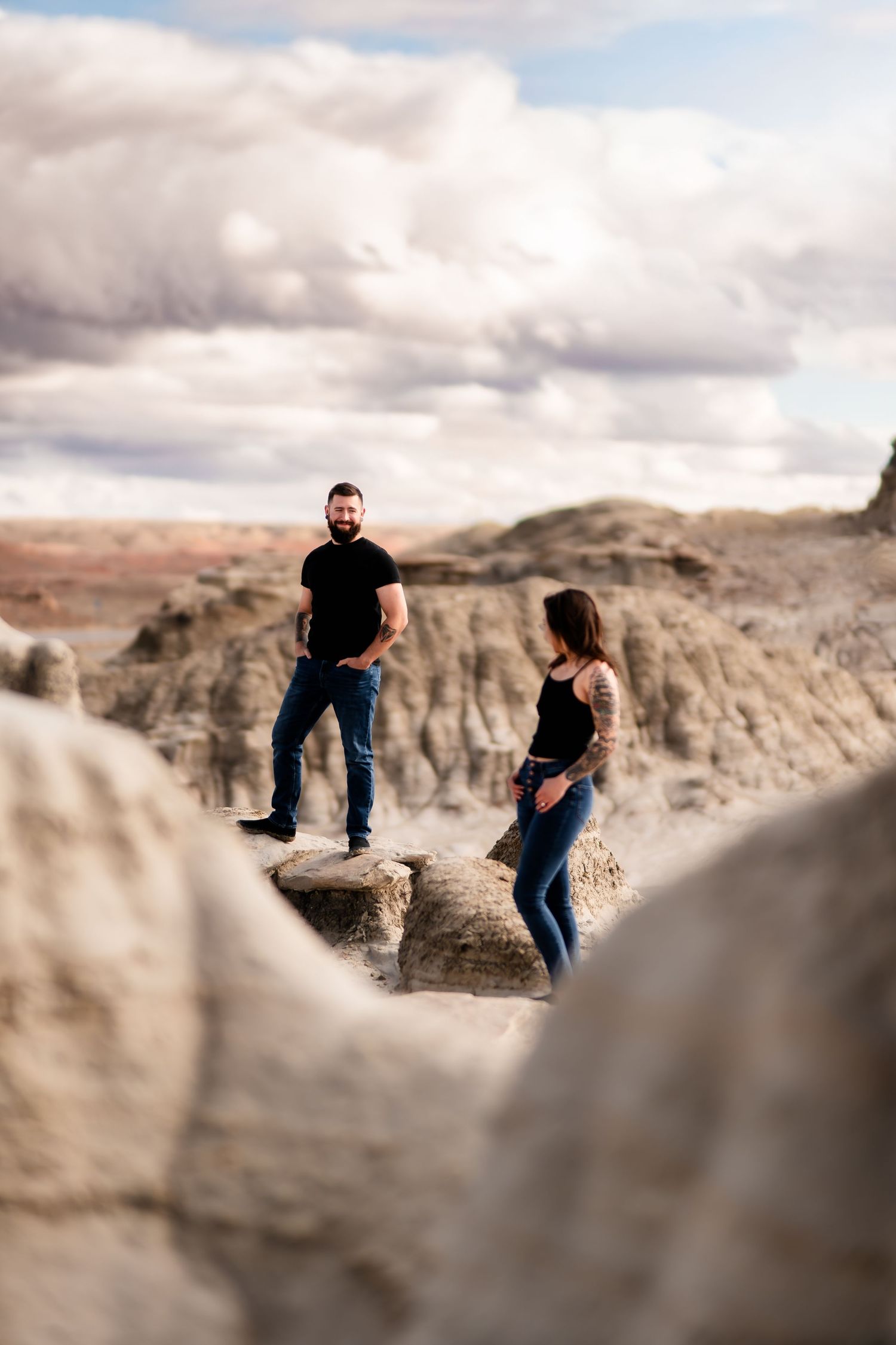 A couple stands on rocky terrain against cloudy skies in a desert landscape with dramatic rock formations.