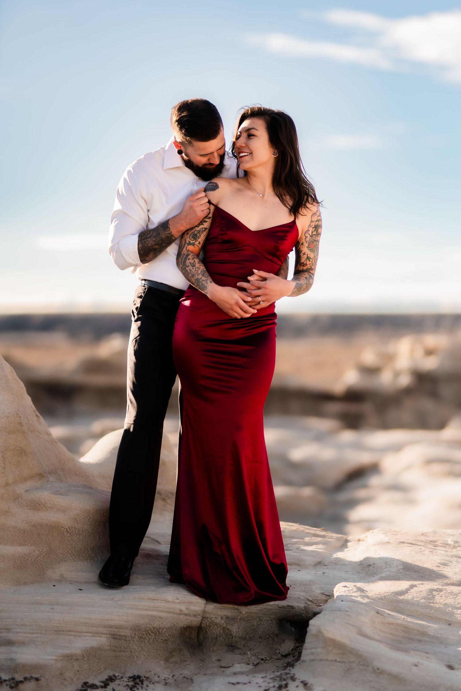 A couple in formal attire embraces on rocky terrain at sunset, with the woman wearing a flowing red velvet dress.