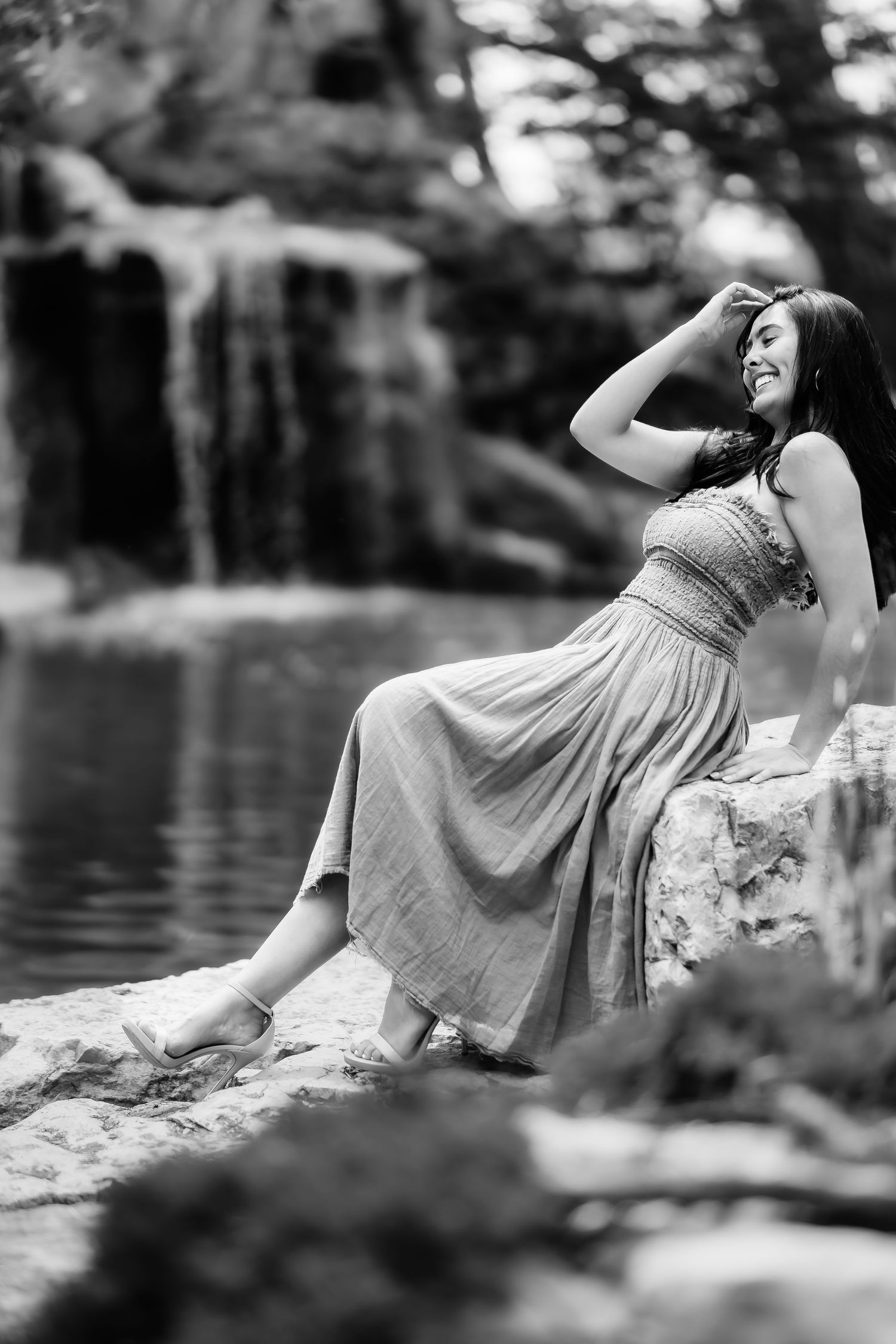 A black and white portrait of someone relaxing on rocks by a serene pond in a flowing dress.