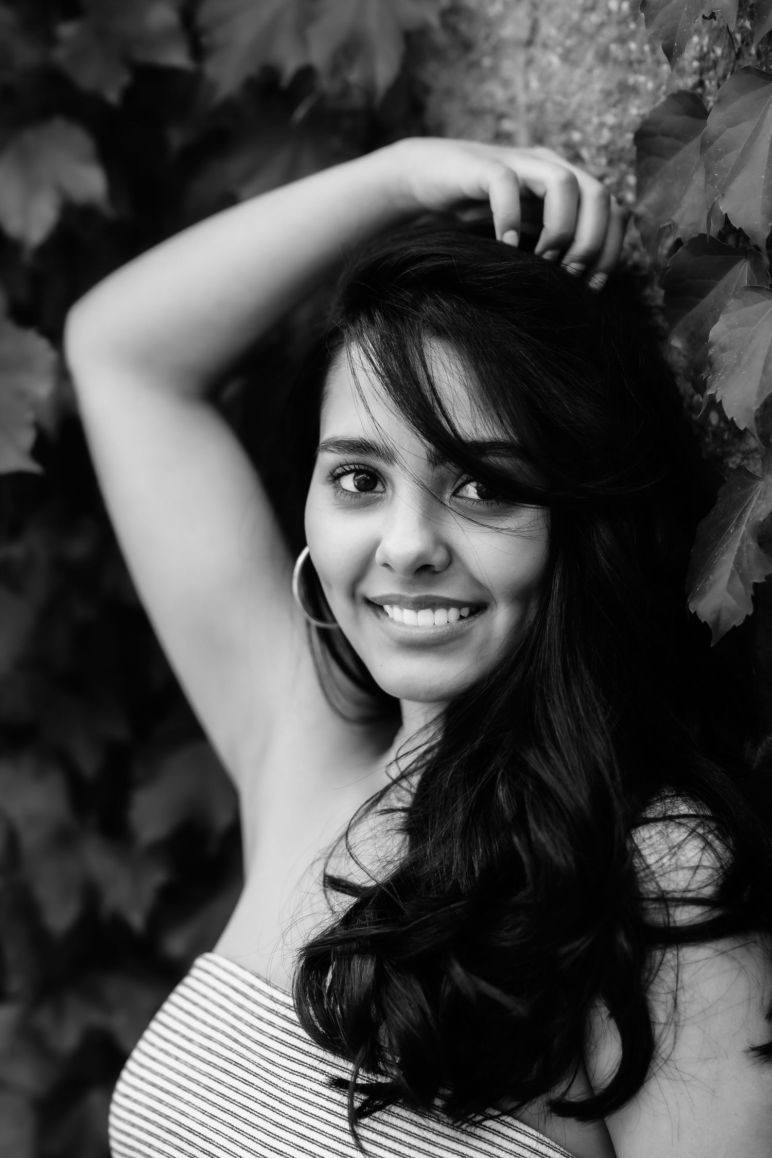 Black and white portrait of someone with dark wavy hair and bright smile wearing a striped top against a leafy background.