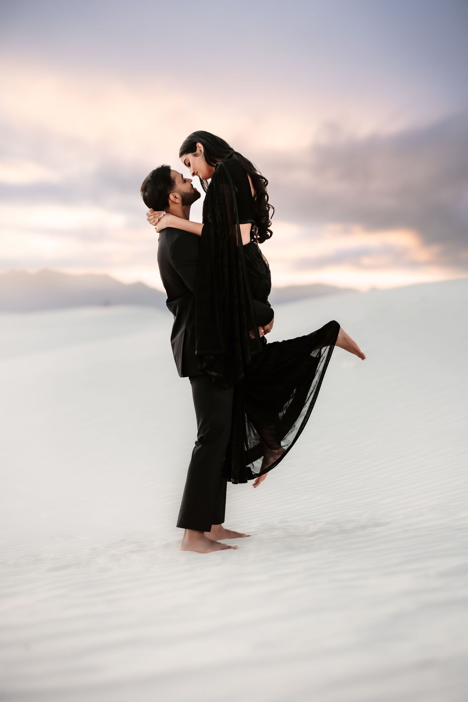 A romantic couple in black outfits embrace while standing on white sand during a dreamy sunset.