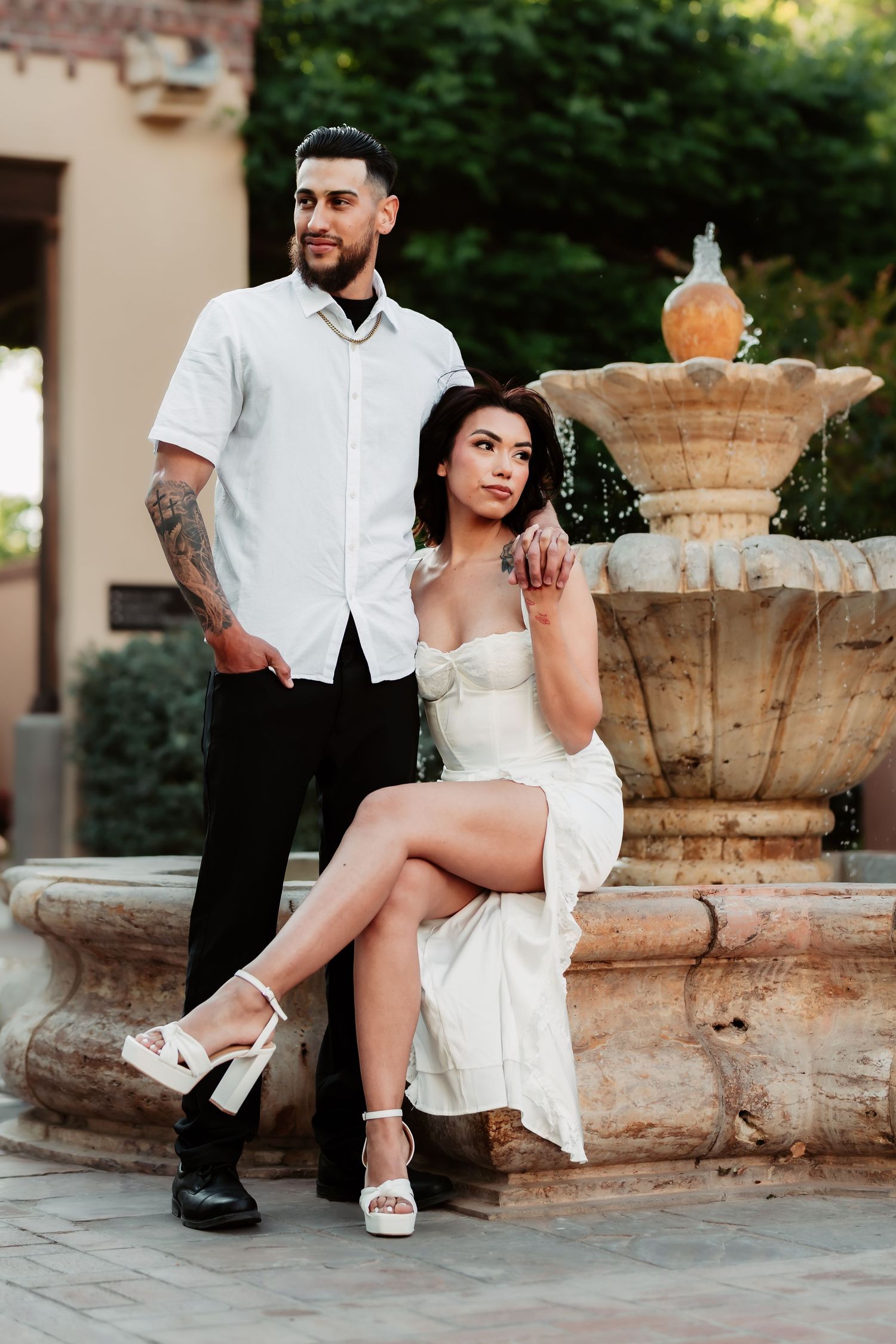 Stylish couple poses by an outdoor fountain wearing coordinated white outfits.