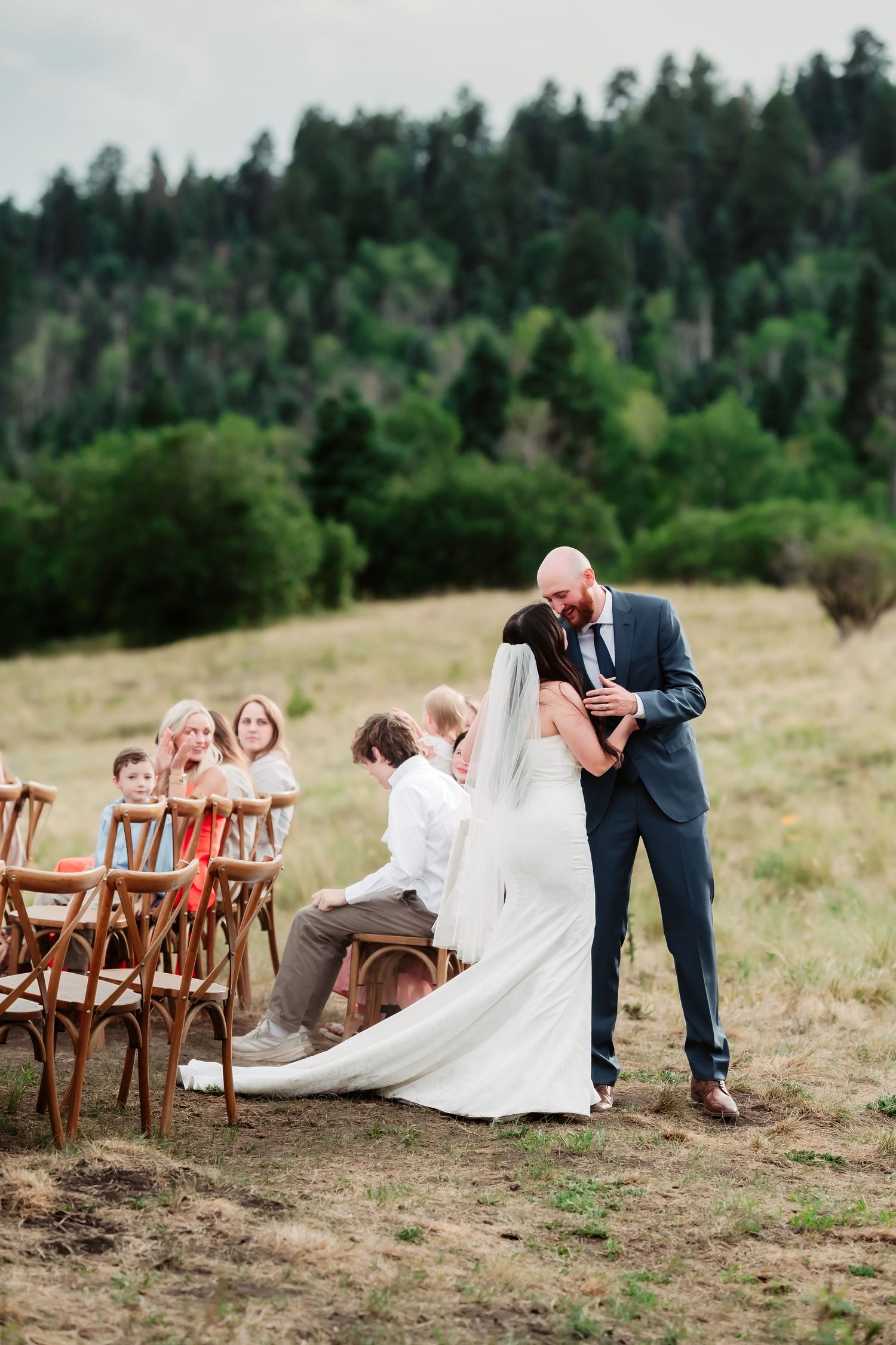 Wedding couple shares intimate moment during ceremony with wooden chairs and forest backdrop.
