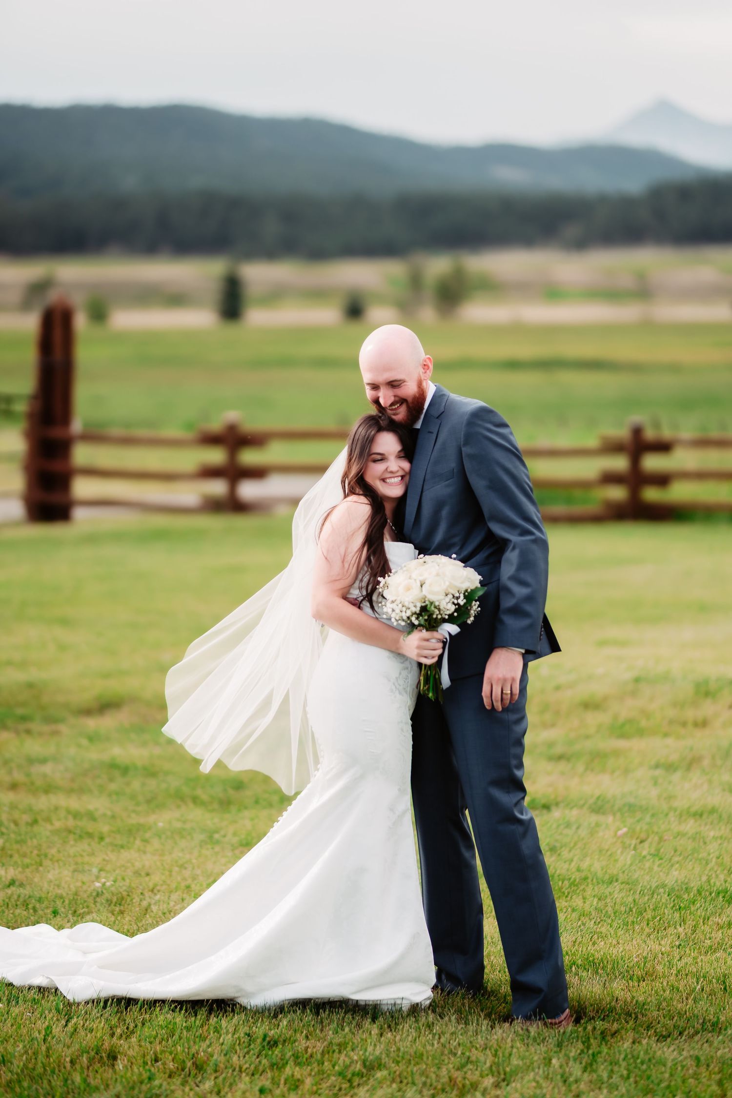 A couple shares playful moments in an outdoor wedding setting with scenic mountain views and rustic fences.
