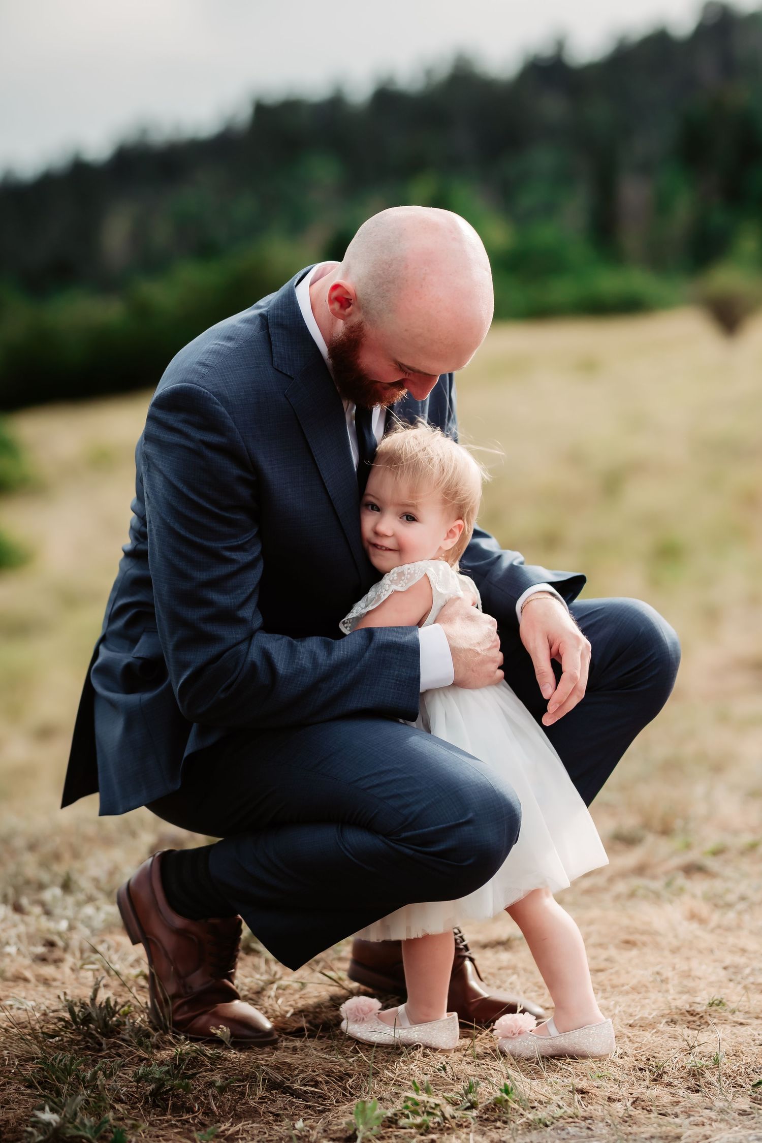 Sweet moment between suited figure and small child in white dress crouching on mountain path.