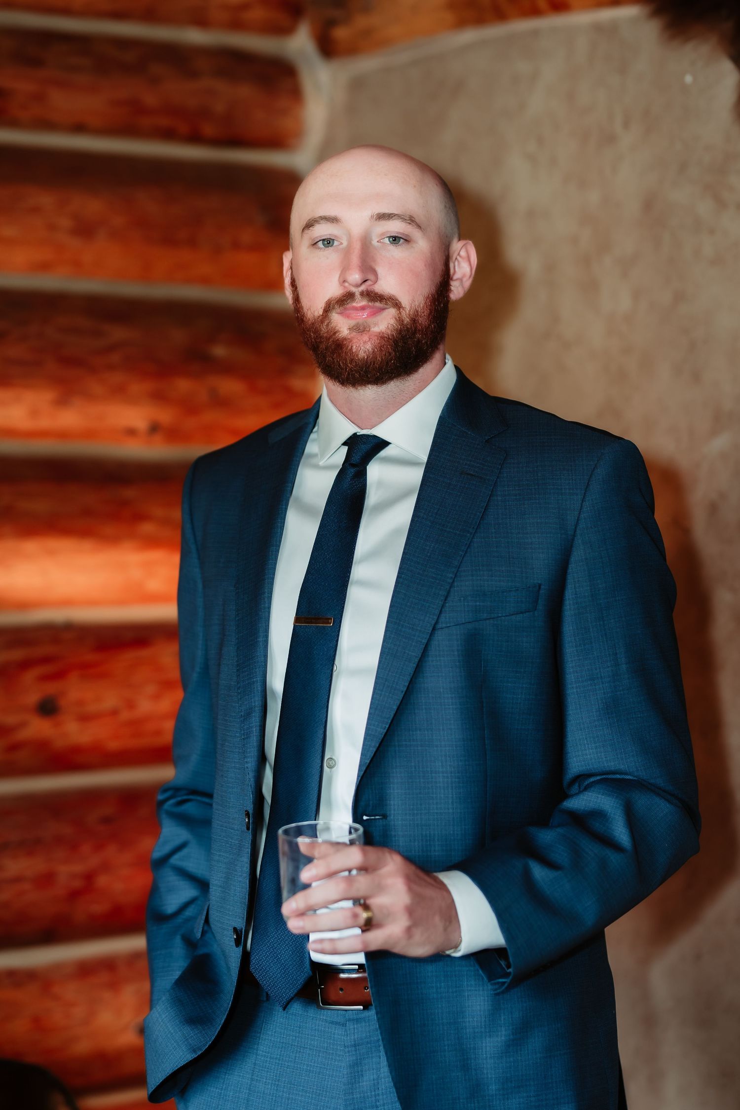 A groom in a blue suit holds a drink while standing against a rustic log cabin wall.