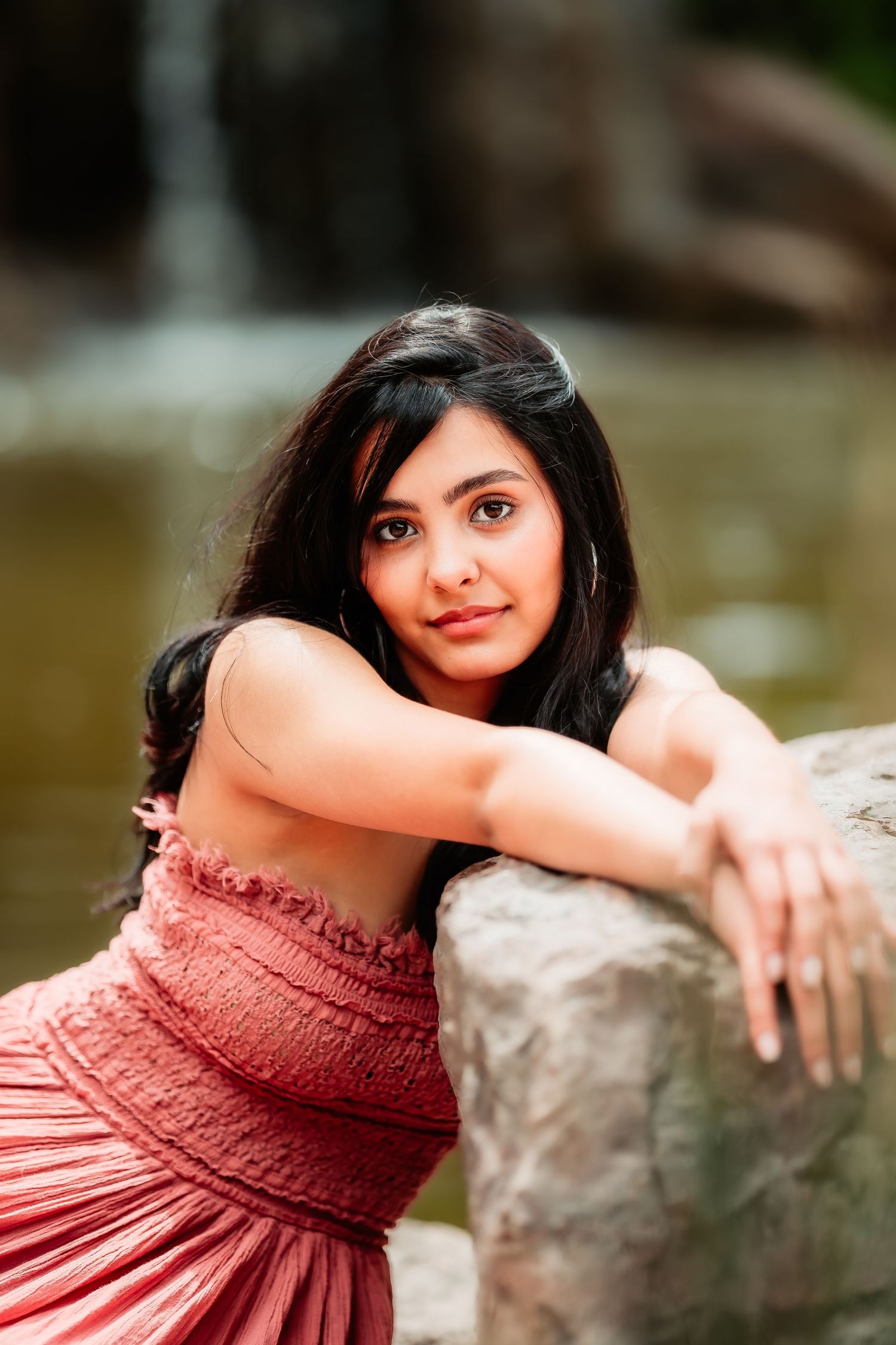 Woman in coral dress leaning against stone wall in outdoor portrait setting.