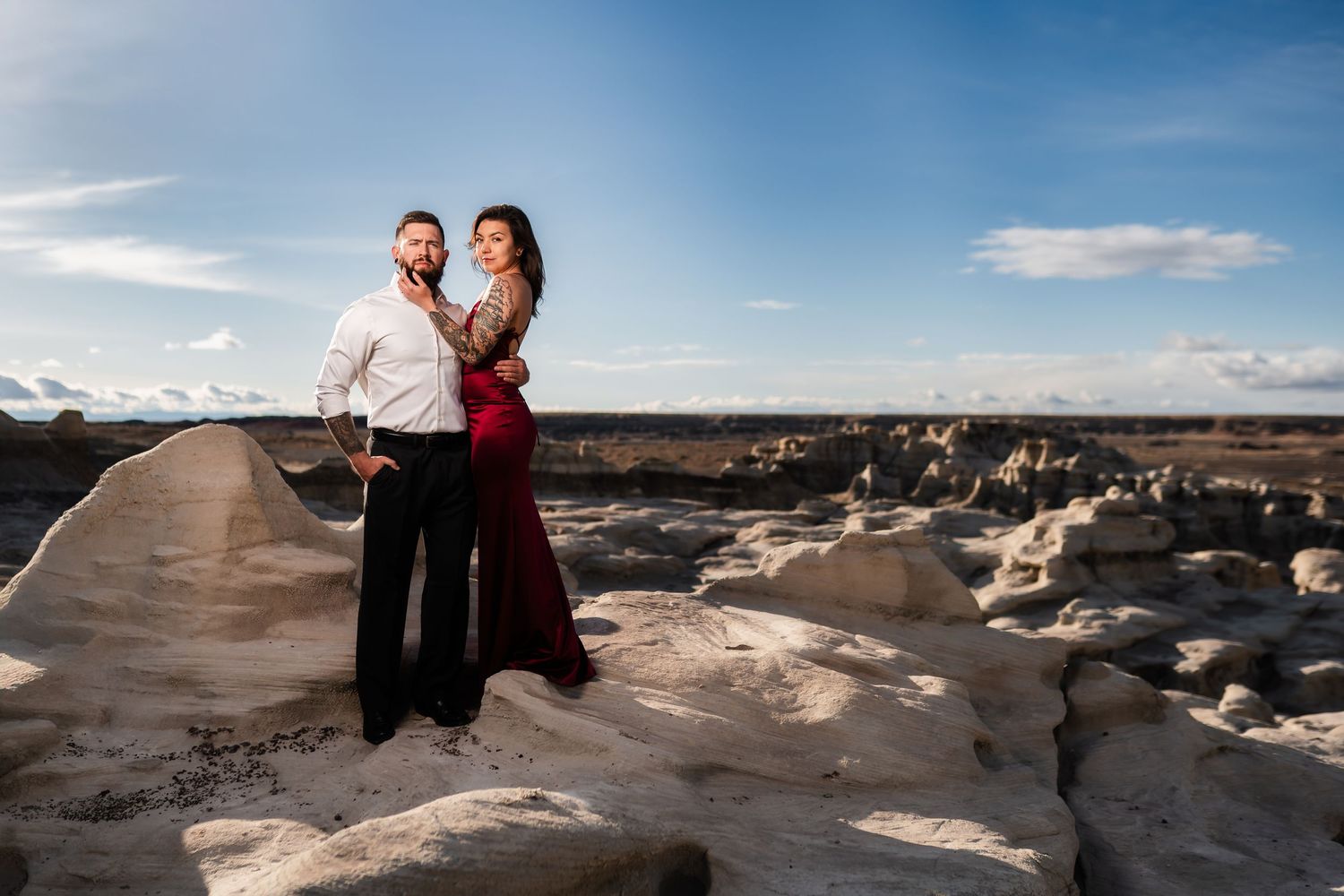 A couple poses together on white rock formations at sunset, with the woman wearing a red dress.