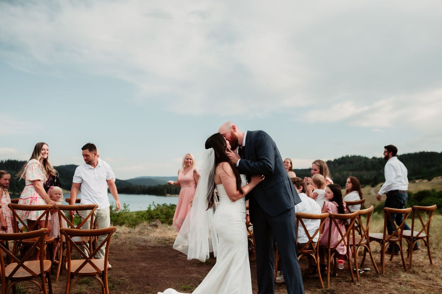 Newlyweds share their first kiss during an outdoor ceremony with wooden cross-back chairs.