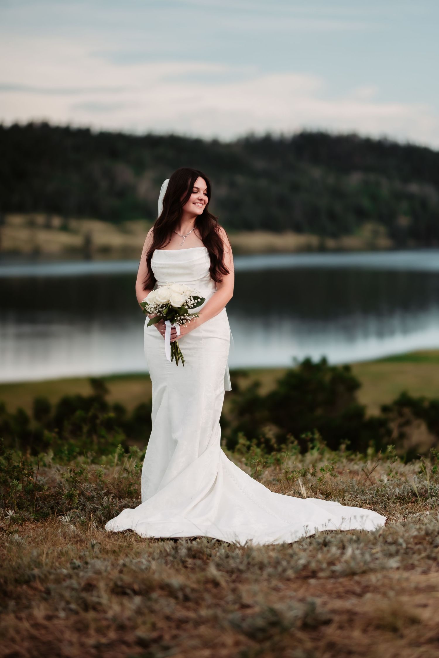 A bride in a white dress holds a bouquet while standing near a serene mountain lake.