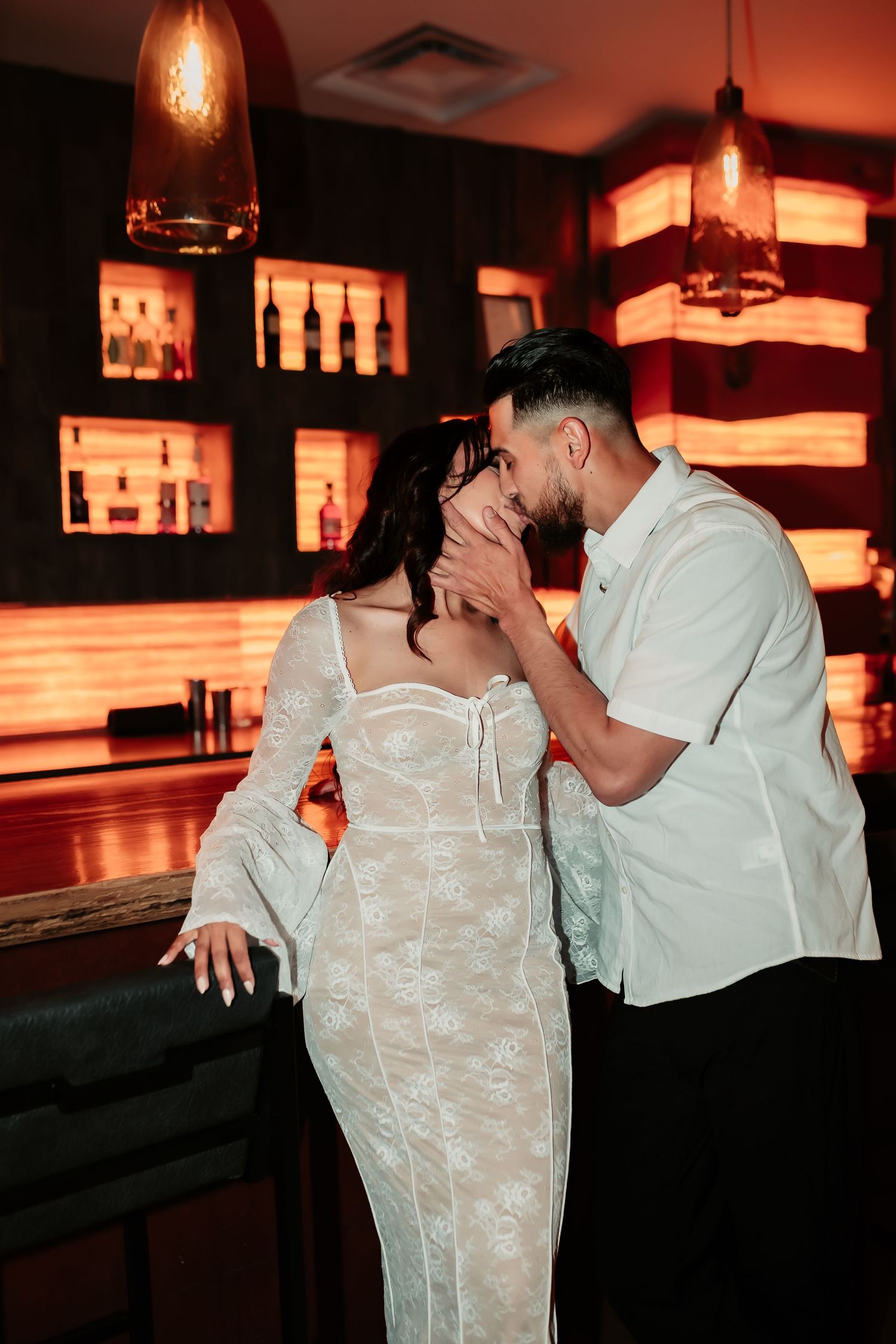 Couple in elegant attire share a romantic moment in a bar with warm ambient lighting and bottle displays.