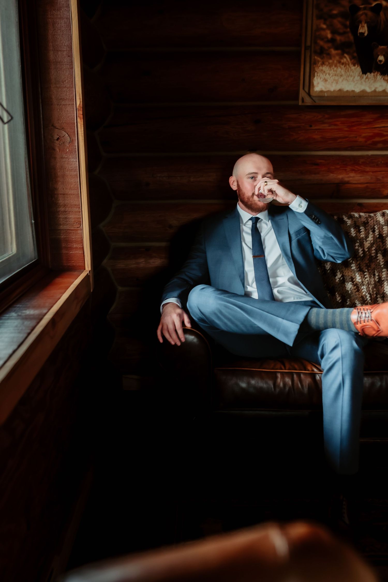 Dramatic portrait in moody lighting showing person seated on leather couch in formal attire.
