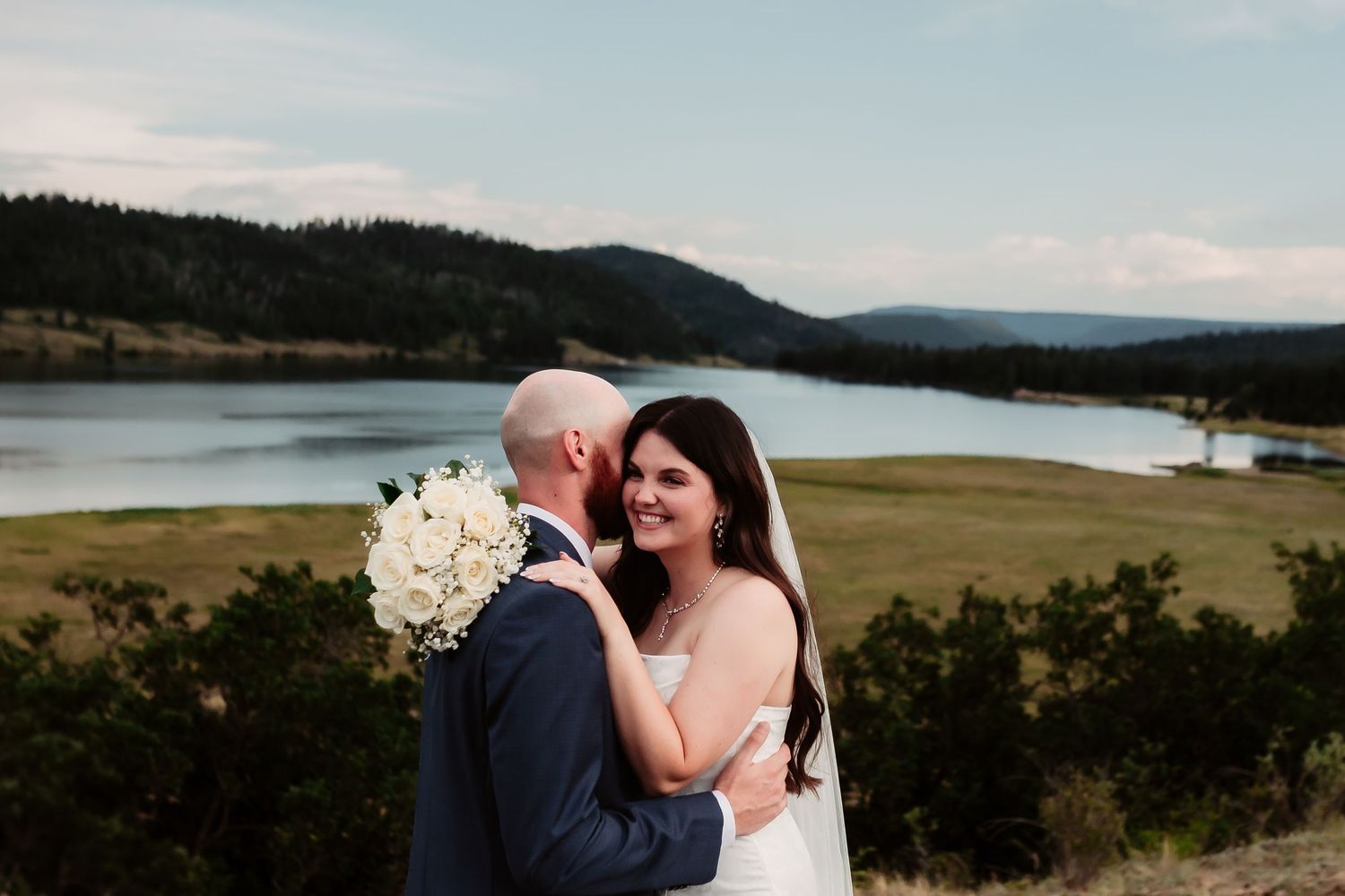 Intimate wedding portrait of couple embracing with scenic lake and mountains in background.