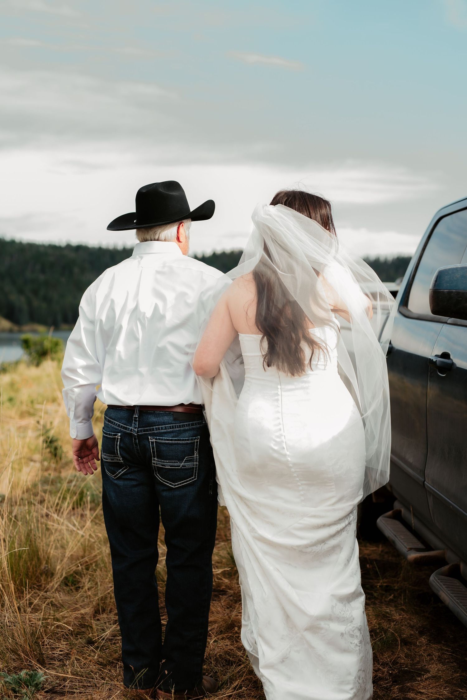 Wedding guests stand against car in scenic mountain setting wearing jeans and formal attire.