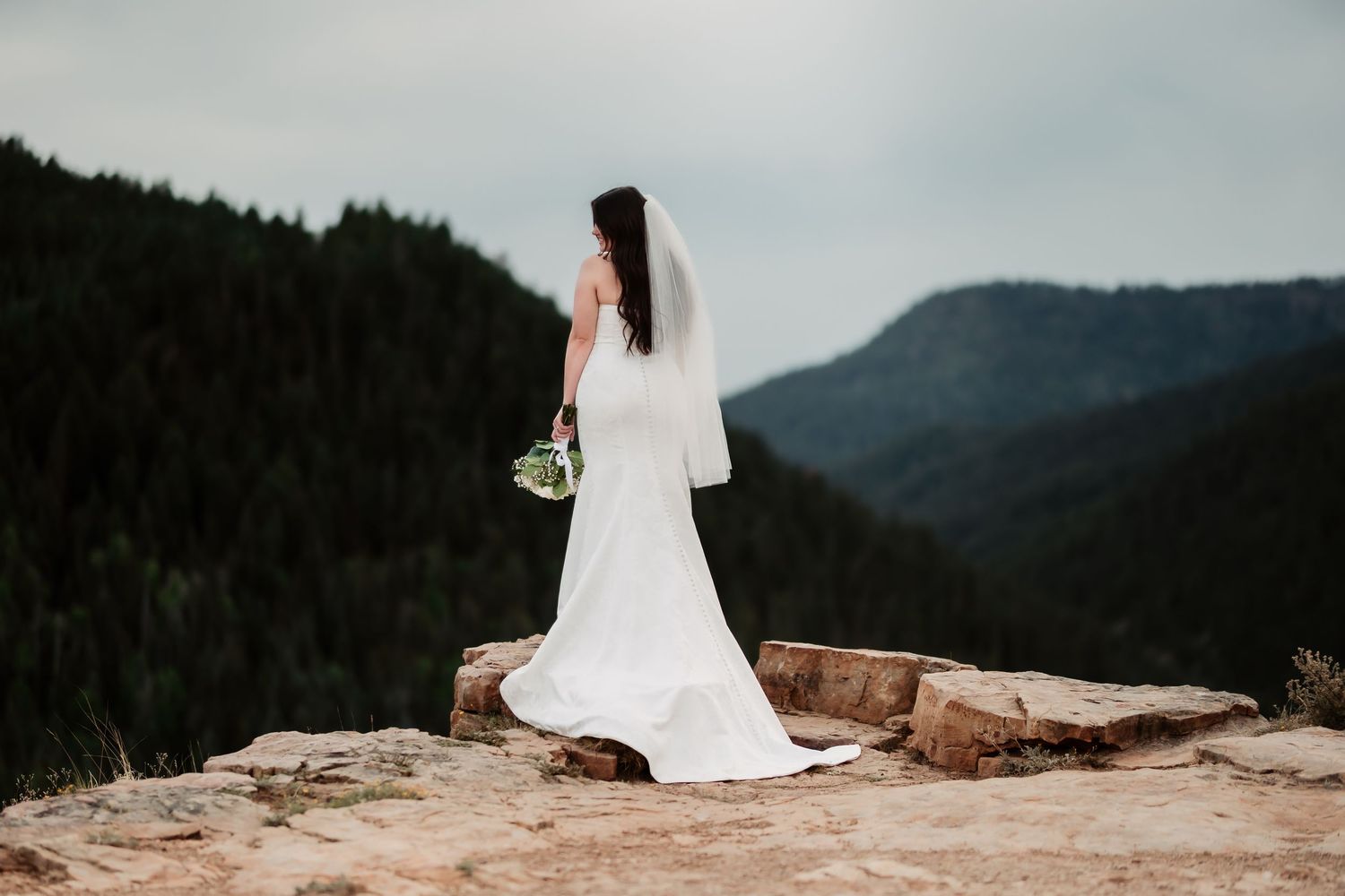 A bride in a flowing white gown stands alone on a mountain ledge holding her bouquet.