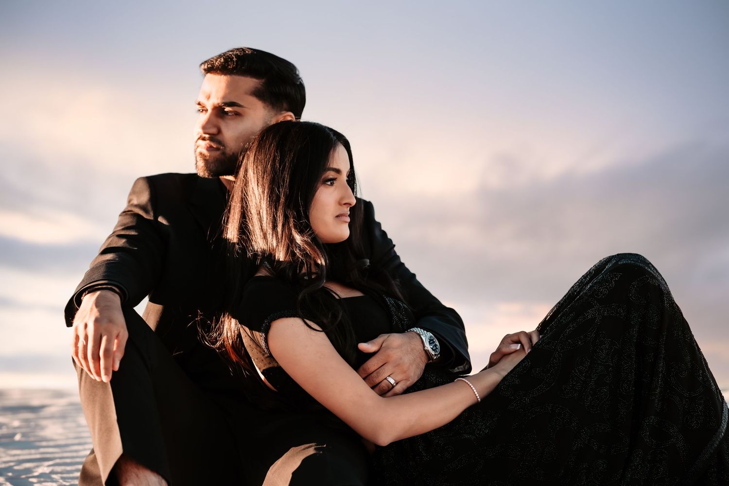 A couple in black outfits poses romantically against a sunset sky during an intimate outdoor photoshoot.