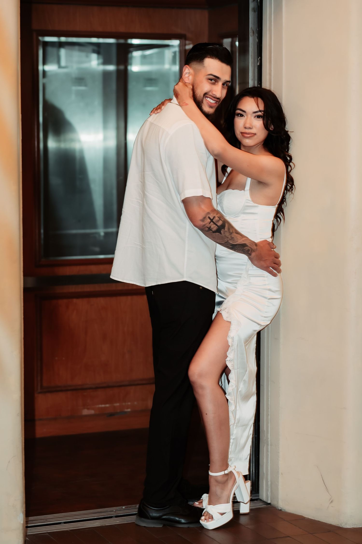 A romantic couple poses intimately in evening attire against a wall near an elevator door.