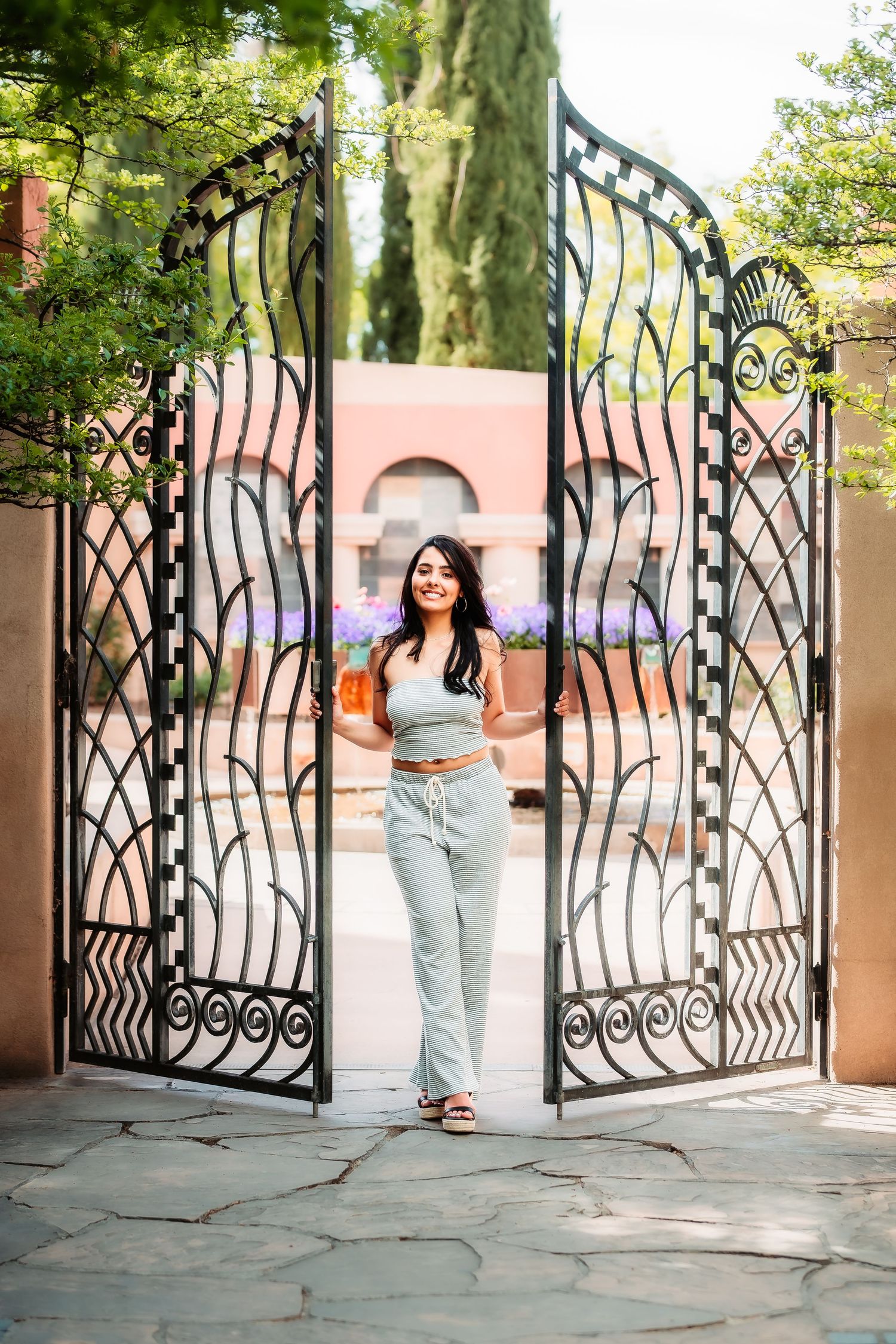 Person standing between ornate iron gates in a garden setting wearing casual summer attire.