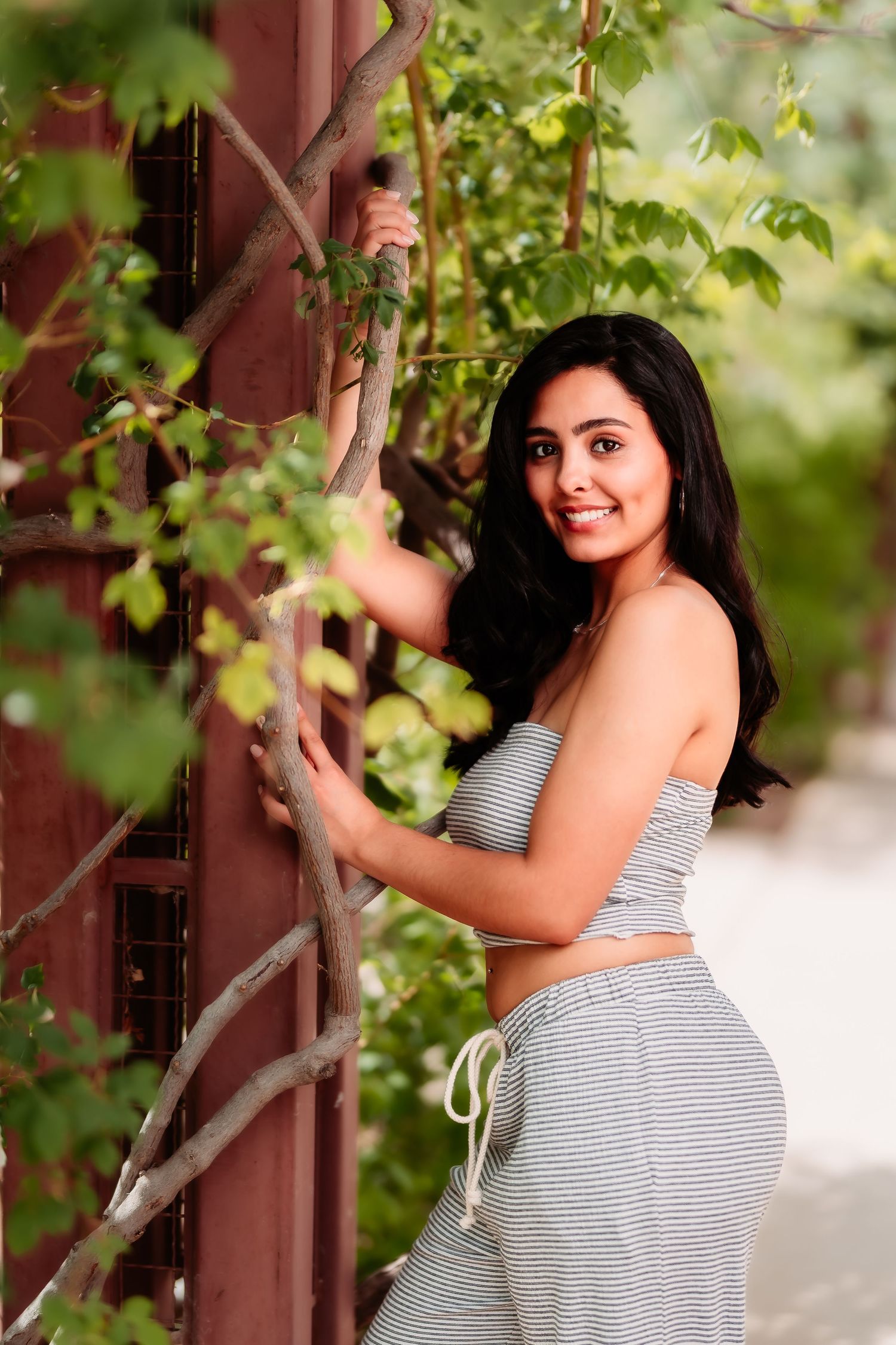 A woman in striped two-piece outfit poses against a garden trellis with green vines and foliage.