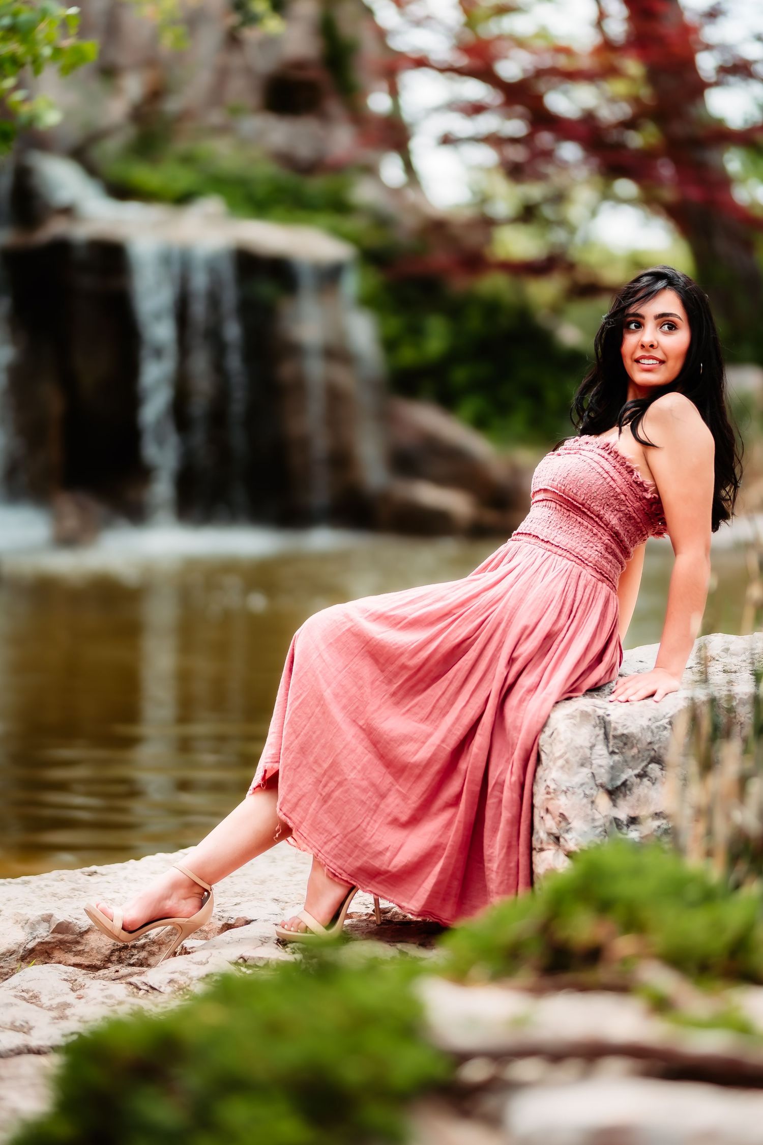 A woman in a coral dress poses elegantly by a waterfall in a garden setting.