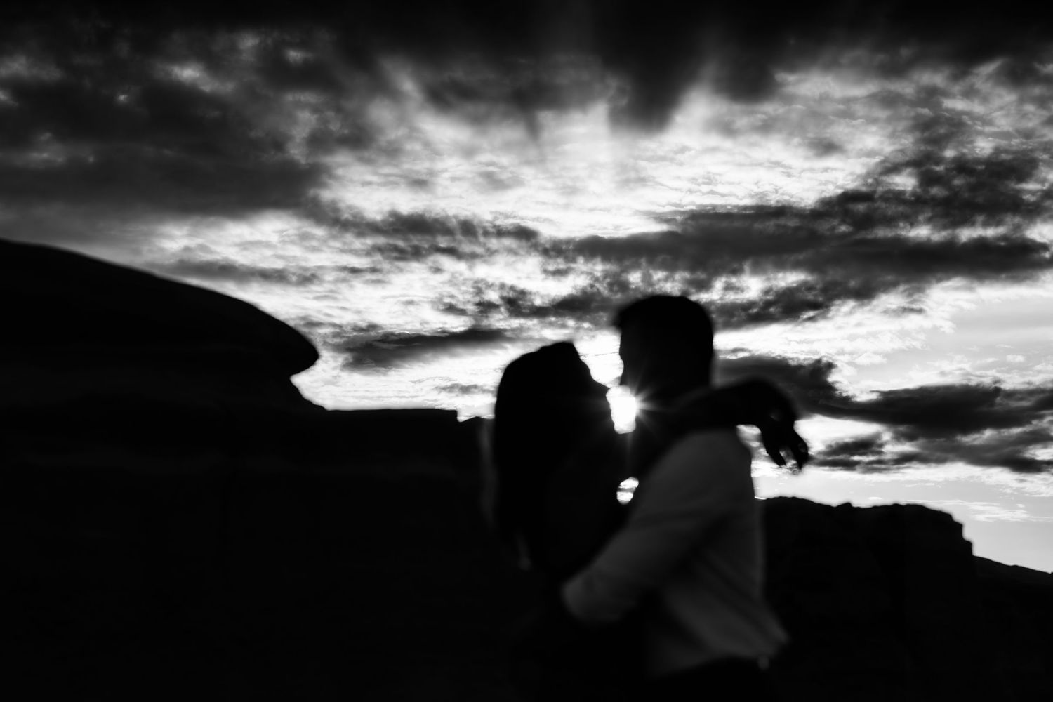 Black and white silhouette of two people standing close together against dramatic cloudy sky and rocky landscape.