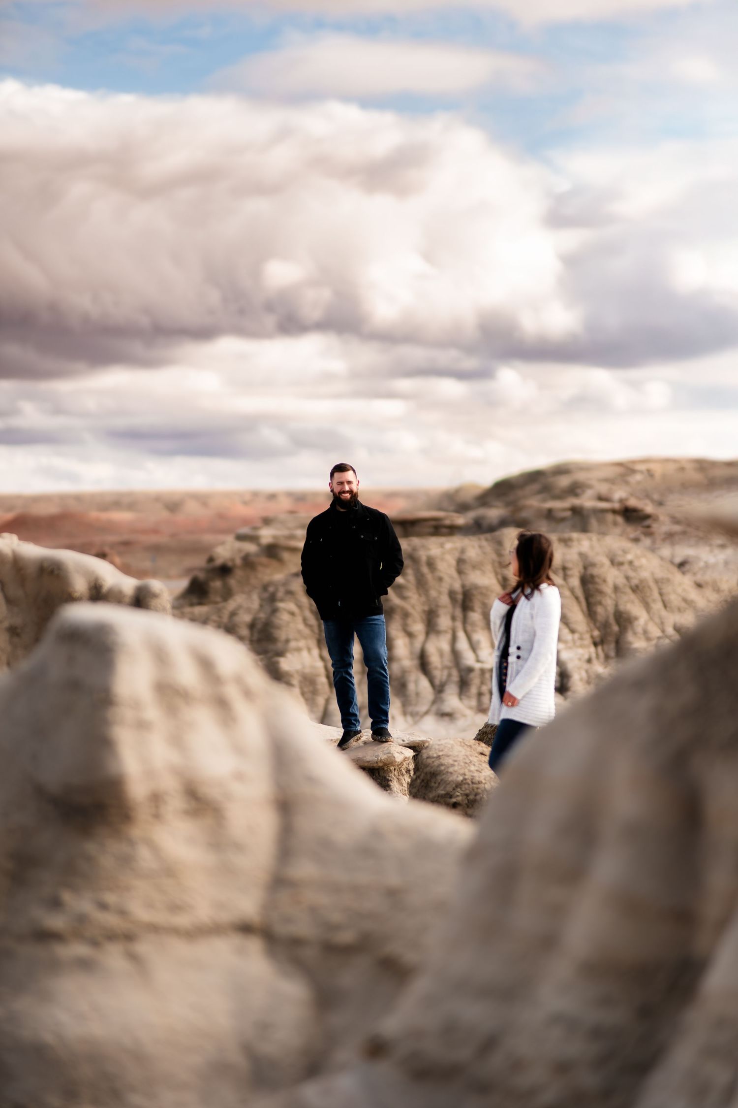 A couple stands on dramatic rock formations against a cloudy sky in a desert landscape.