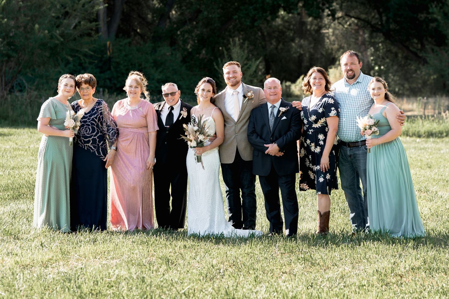 Wedding party group photo with bridesmaids and groomsmen in pastel dresses and dark suits outdoors.