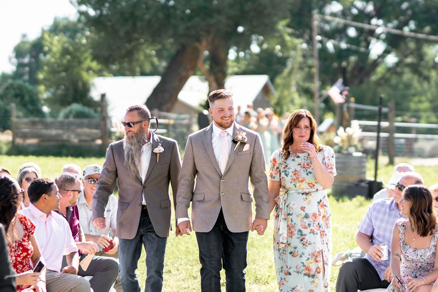 Wedding attendees walk down an outdoor aisle lined with guests on a sunny summer day.