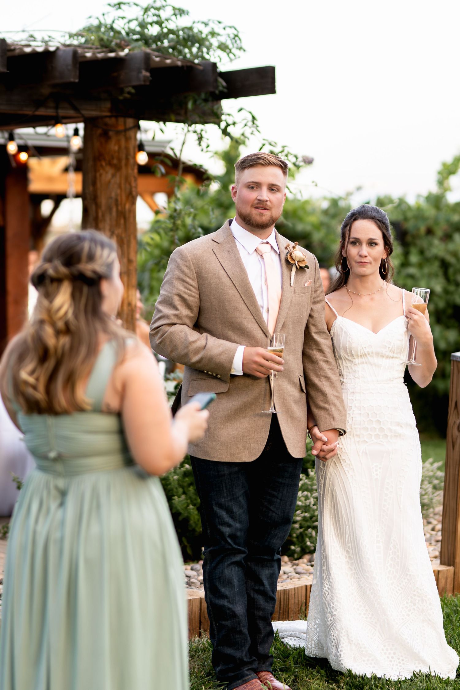 A wedding couple stands under a rustic wooden pergola during their outdoor ceremony.