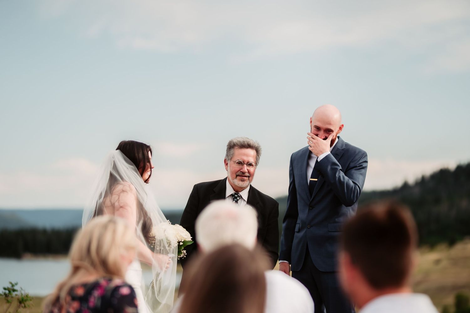 An outdoor wedding ceremony takes place with a scenic lake and mountains in the background.