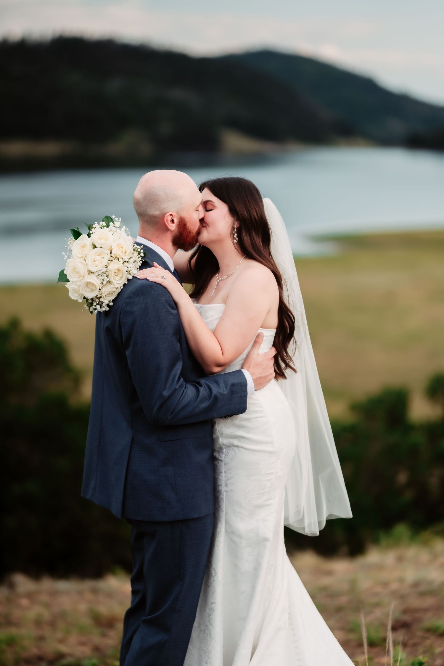 Newlywed couple sharing a romantic kiss by a scenic mountain lake, with the bride holding a white floral bouquet.