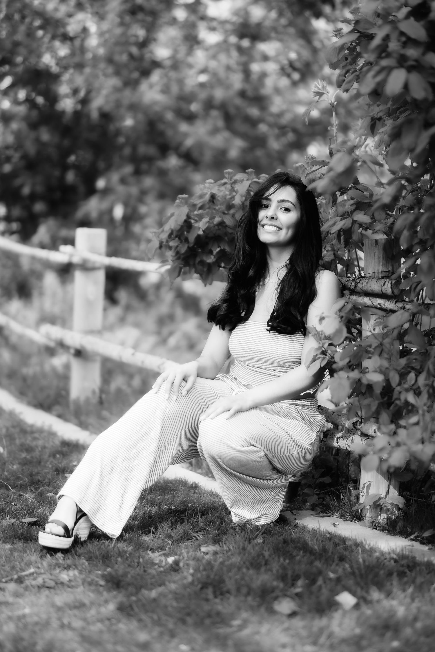 Black and white outdoor portrait near a rustic fence and foliage wearing a light summer outfit.