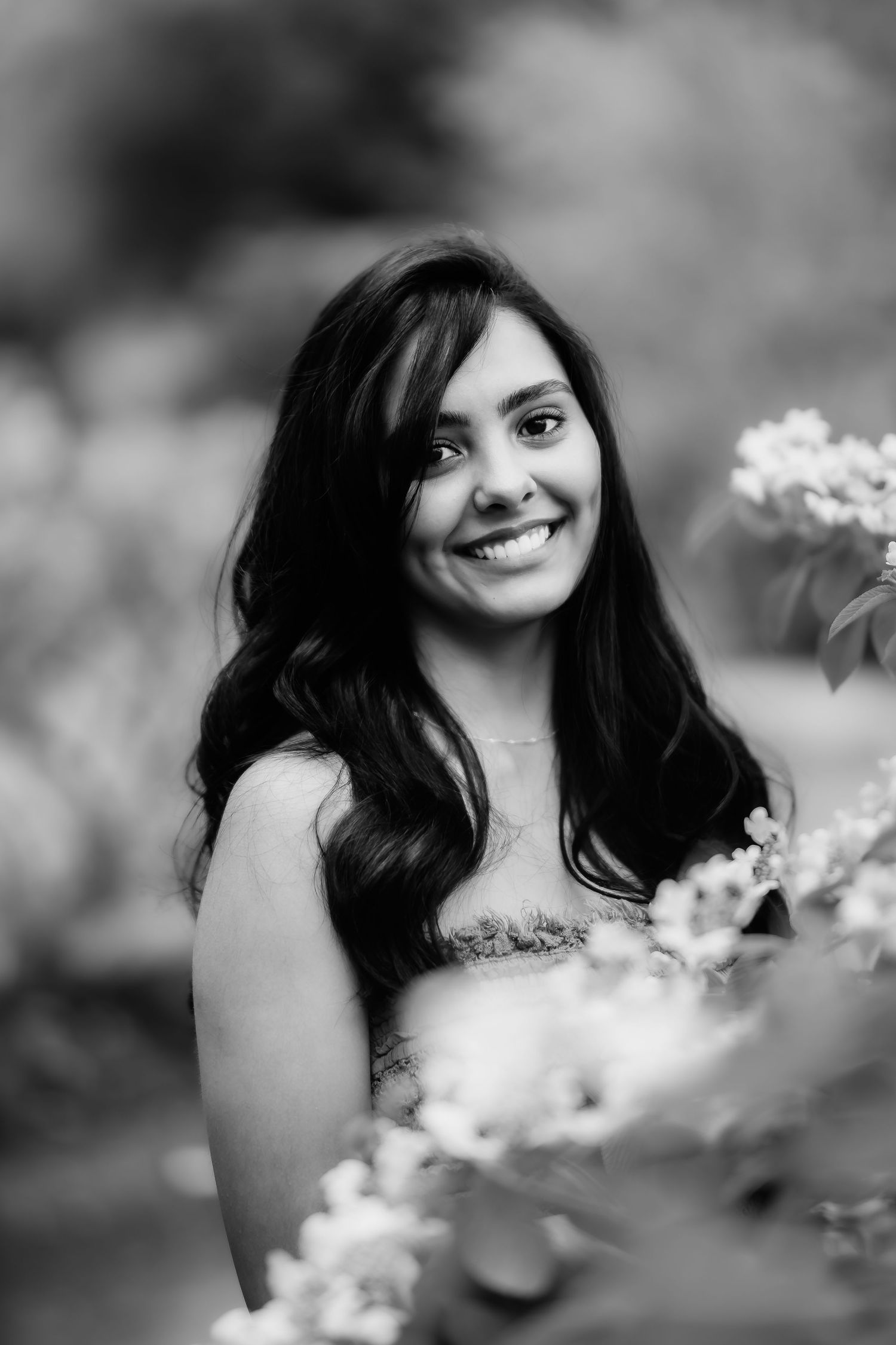 Black and white portrait of woman with long dark hair smiling among flowers in outdoor setting.