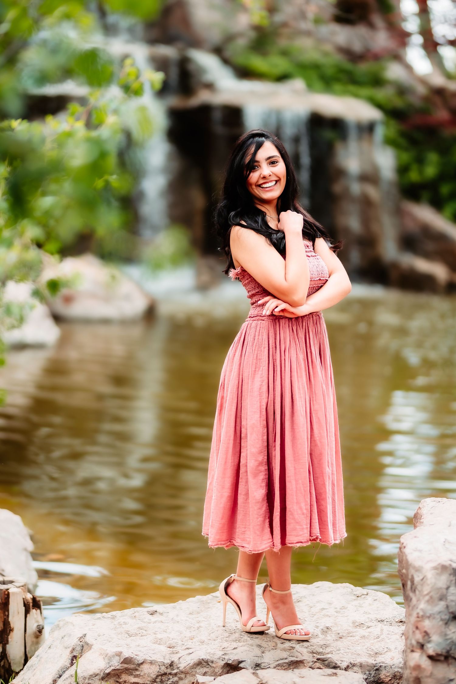 Woman in coral pink dress standing gracefully by a waterfall and pond in an outdoor garden setting.