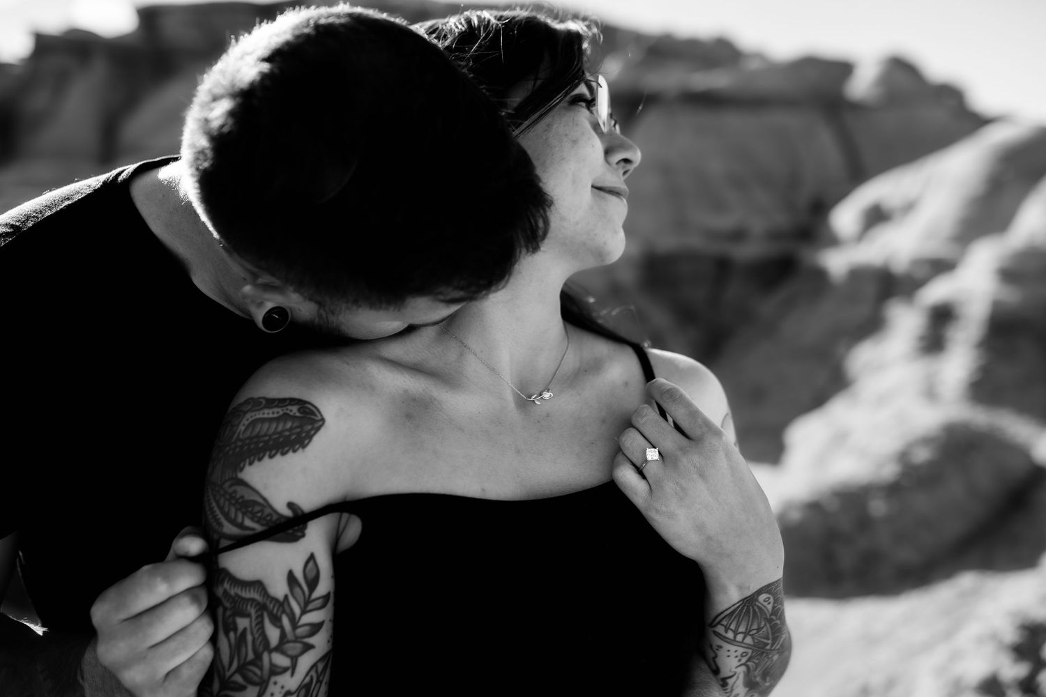 An intimate black and white portrait of two people embracing outdoors with visible tattoos.