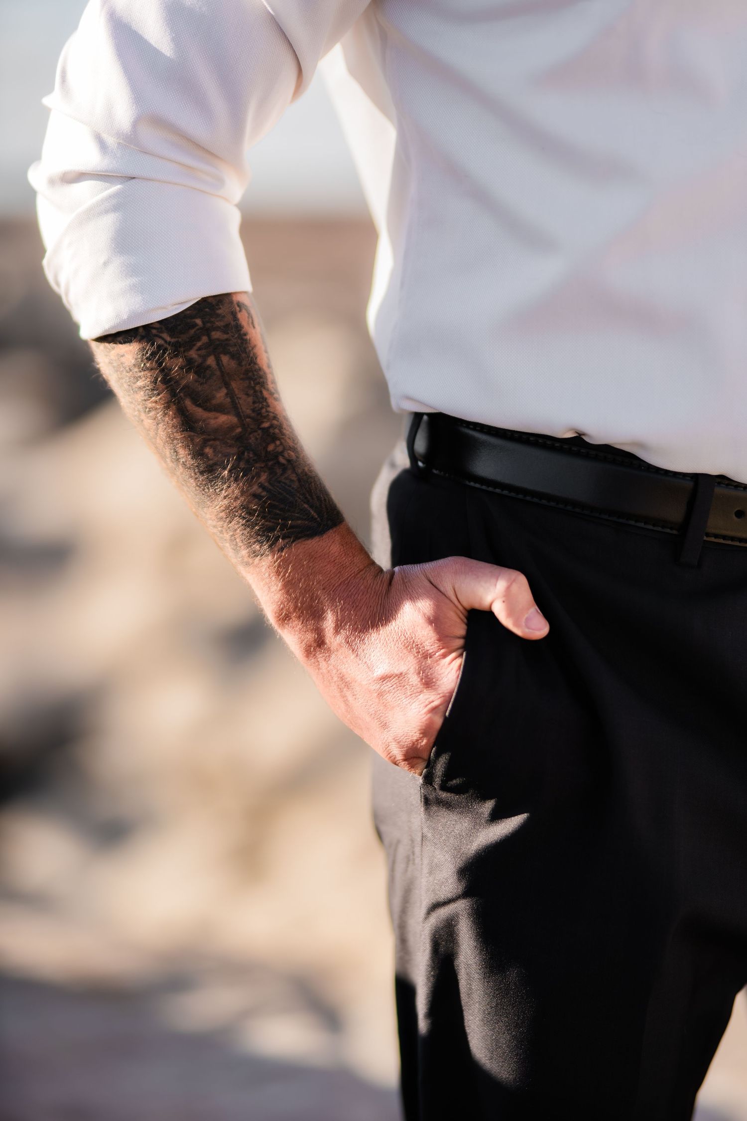 Close-up of a tattooed arm in white shirt sleeve with hand resting on hip.