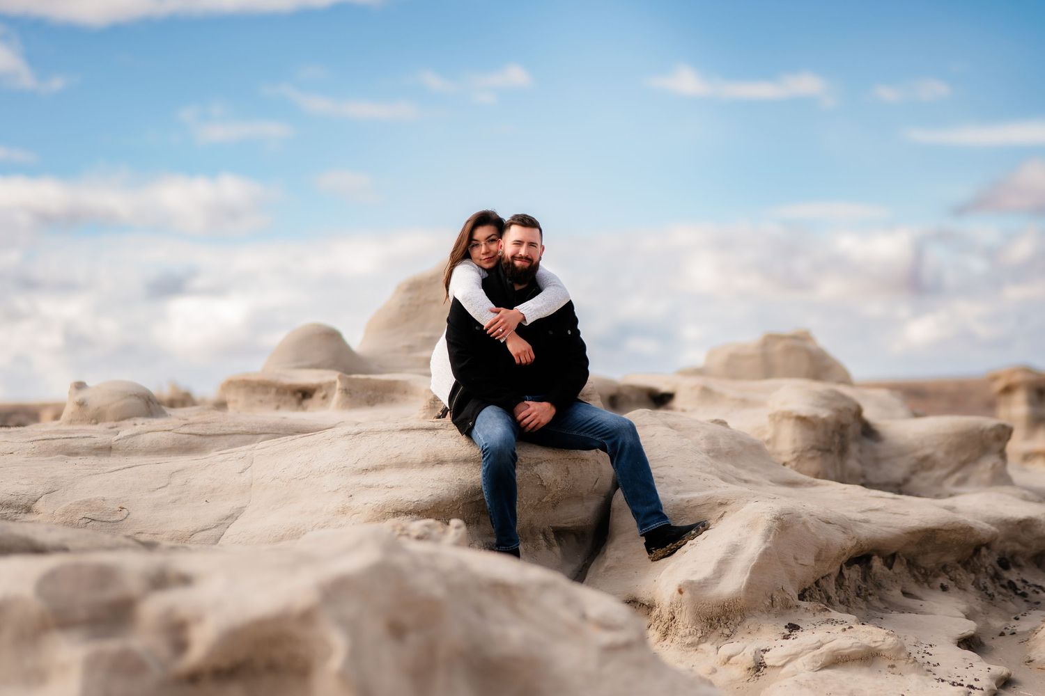 A couple embraces while sitting on weathered desert rock formations on a bright sunny day.