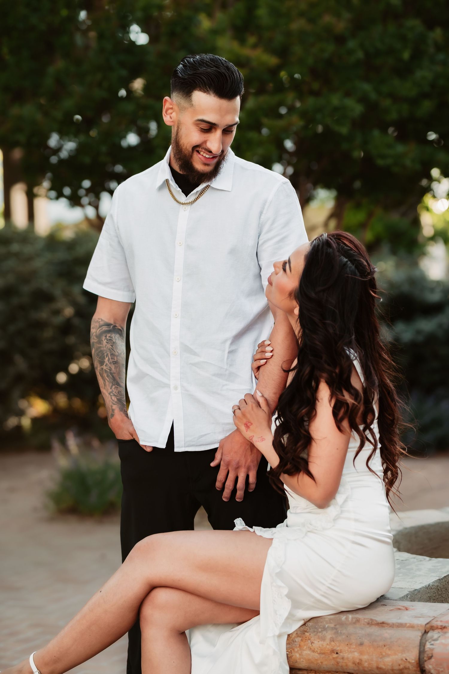 Romantic couple in coordinating white outfits share a tender moment outdoors near garden landscaping.