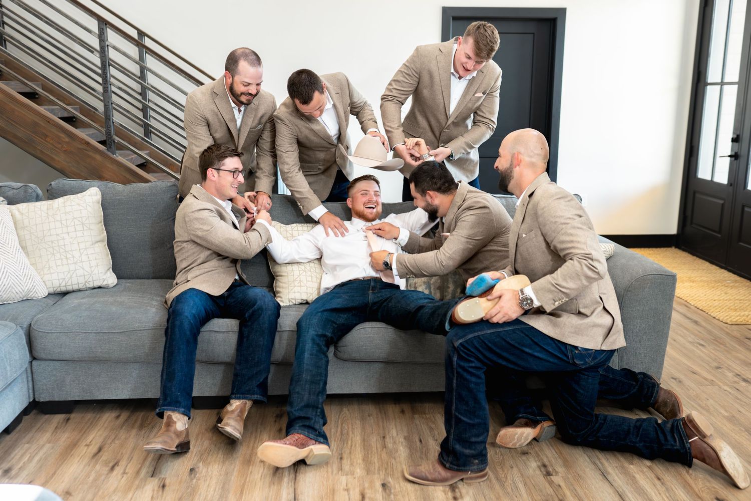 Groomsmen in beige suits relaxing and laughing together on a gray couch in modern interior.