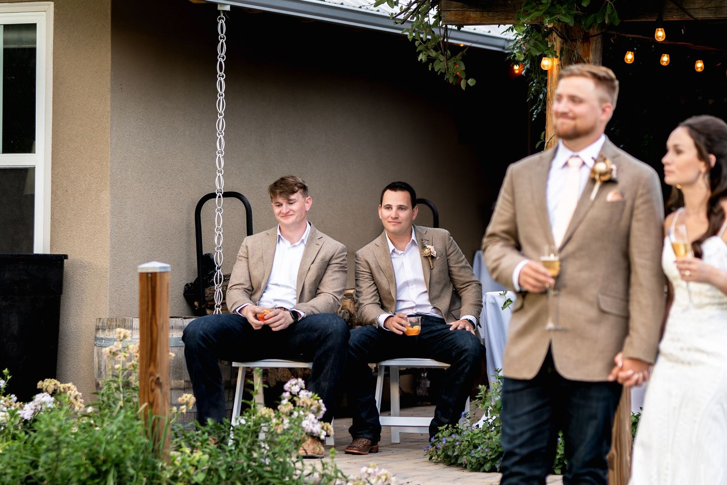 Wedding guests relax on porch swing while couple walks past.